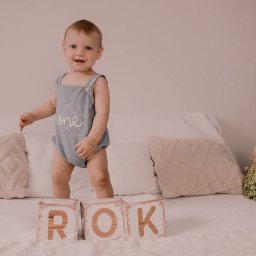 Fotografia Rodzinna Irina Kazarinova - Radosny roczny chłopiec w niebieskim stroju z napisem 'one' stoi na łóżku obok drewnianych klocków z napisem 'ROK', kwiaty w tle, ciepłe oświetlenie.