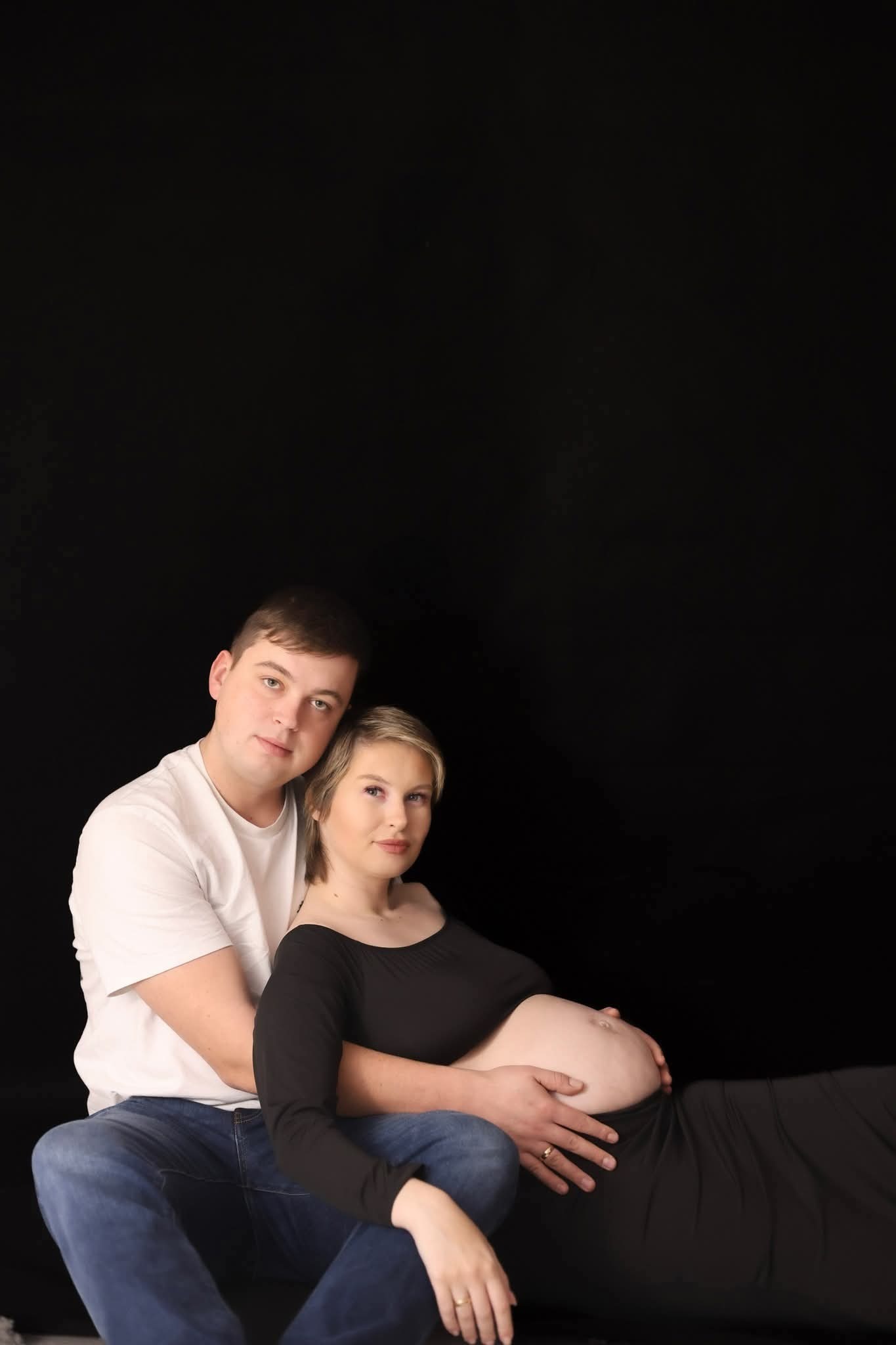 Czułe ujęcie pary: mężczyzna obejmuje brzemienną kobietę w czarnej sukni na ciemnym tle. Dłonie na brzuchu, subtelne światło podkreśla intymność sesji.