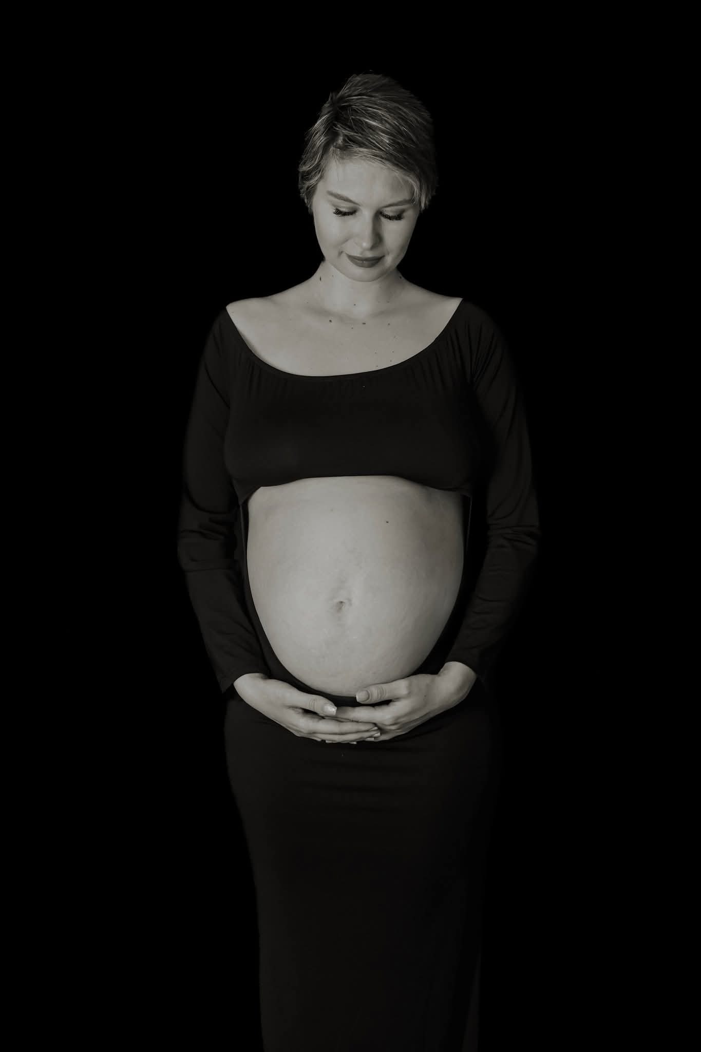 Monochromatyczne ujęcie kobiety w ciąży, dłonie na brzuchu, wzrok skierowany w dół, ciemne tło, subtelne światło podkreśla kształty, delikatny uśmiech.
