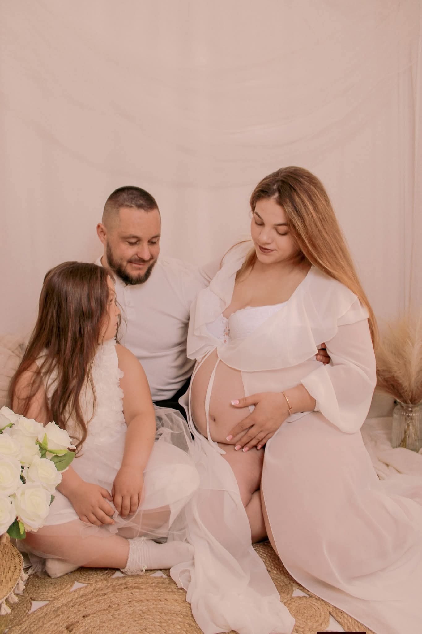 Uśmiechnięta, przyszła mama w bieli, z partnerem i córką, pozuje na jasnym tle z kwiatami. Delikatne światło podkreśla intymny charakter sesji ciążowej.