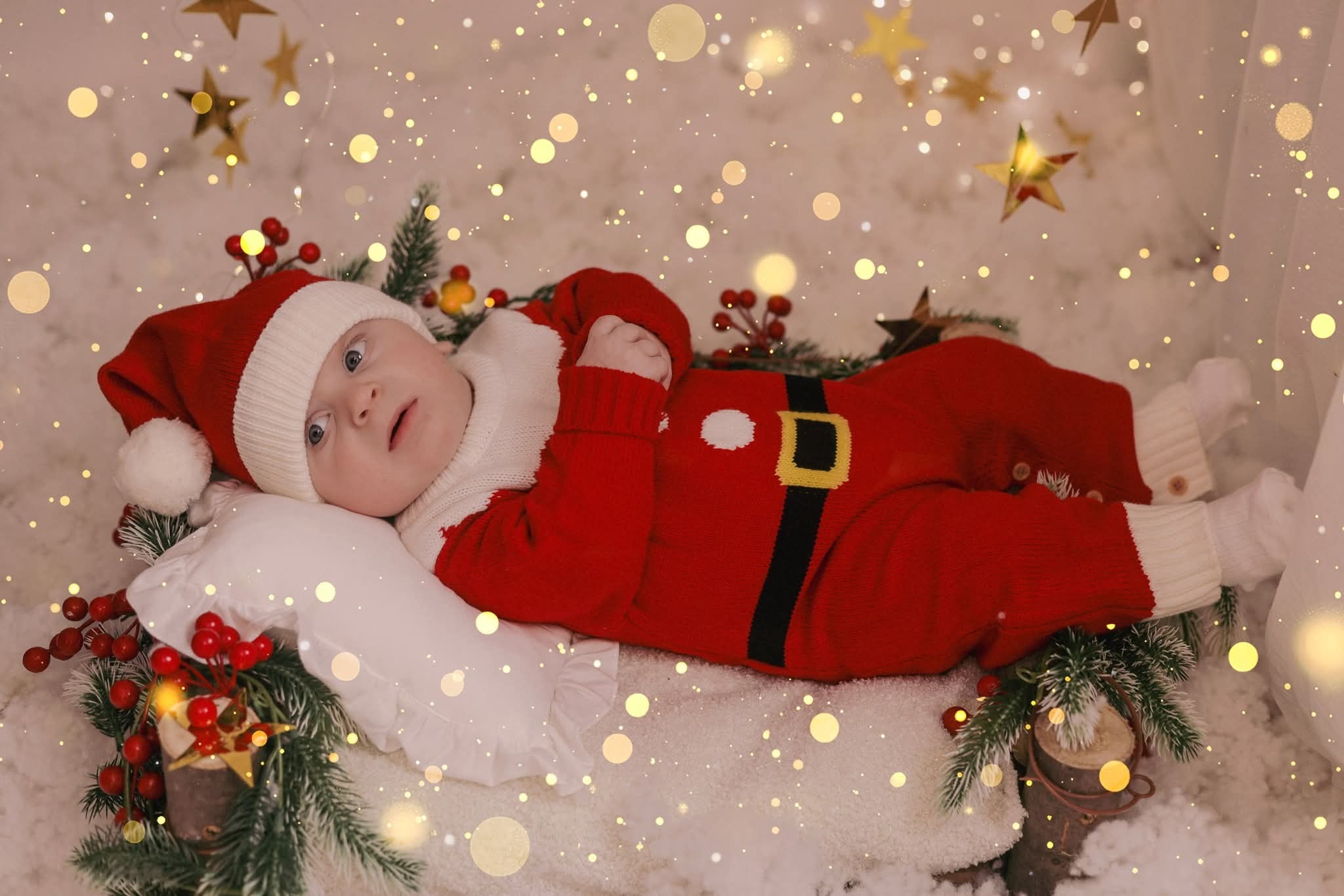 Niemowlę w czerwonym stroju Mikołaja leży na białym kocyku, otoczone świątecznymi dekoracjami i delikatnymi światełkami, tworząc ciepłą, świąteczną atmosferę.