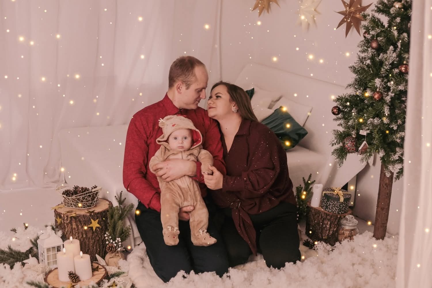 Rodzinne zdjęcie w świątecznej scenerii: rodzice patrzą na siebie, trzymając ubrane w renifera dziecko na tle choinki i dekoracji.