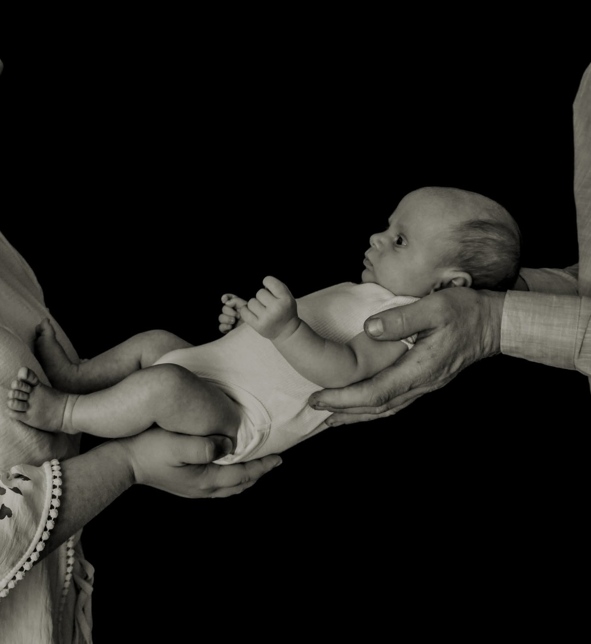 Czarno-białe zdjęcie noworodka w białym body, trzymanego w dłoniach rodziców na tle czerni. Ujęcie z profilu, skupione na twarzy dziecka. Delikatne światło podkreśla rysy.
