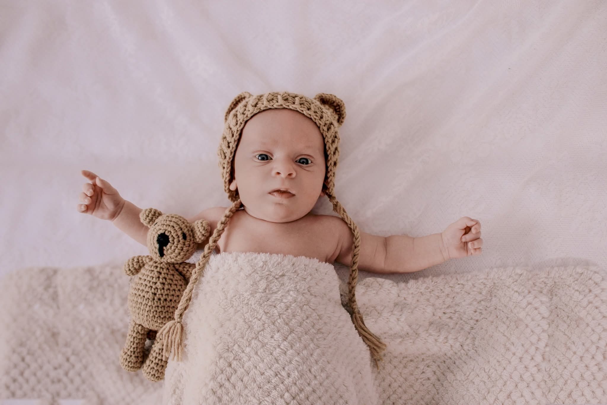 Niemowlę w beżowym, dzierganym kapeluszu z uszkami misia, leżące na puszystym kocyku z pluszowym misiem obok, ujęcie z góry, delikatne światło.