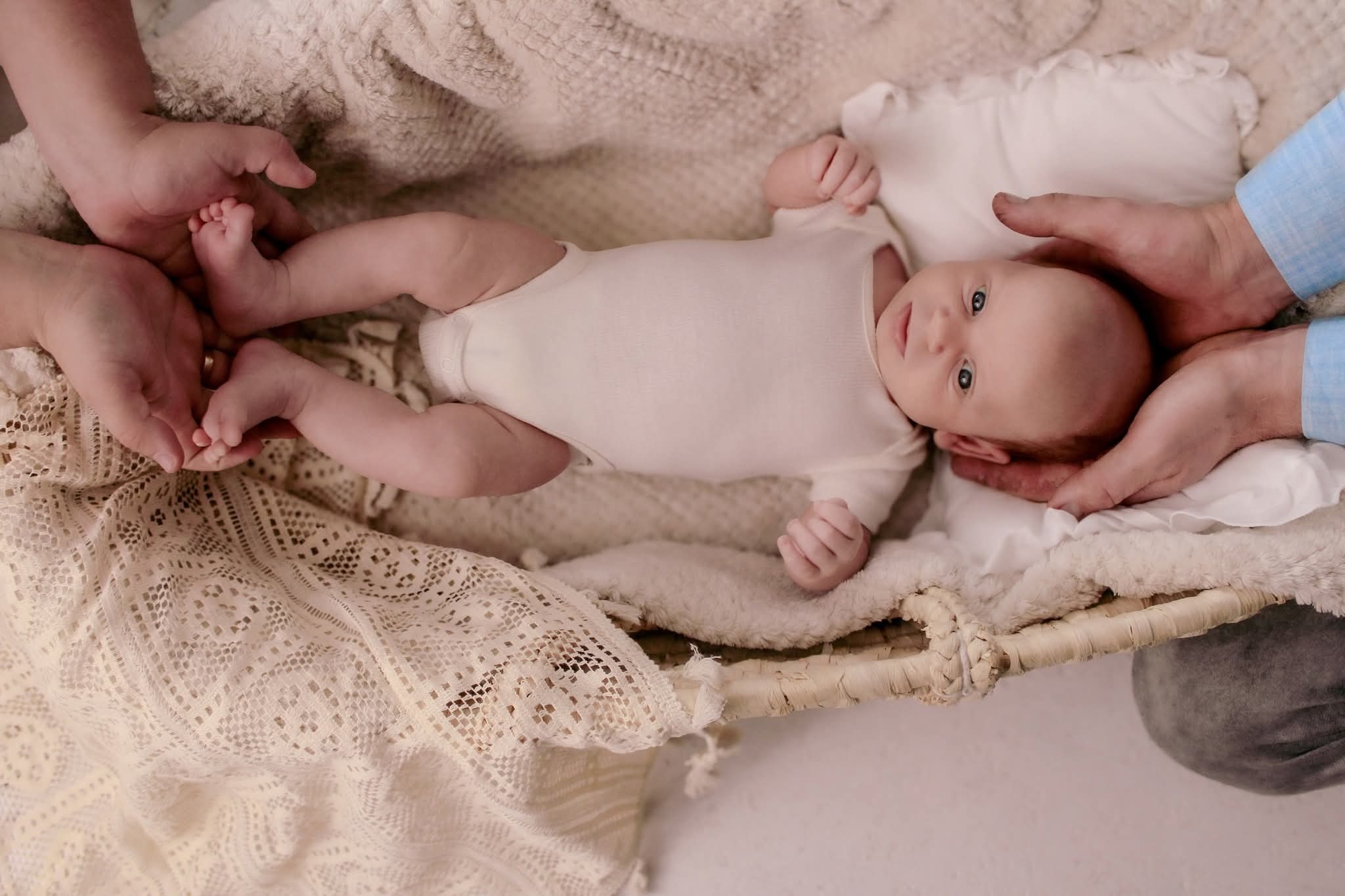 Niemowlę w białym body leży w koszyku, otoczone dłońmi rodziców. Delikatne, naturalne światło podkreśla niewinność dziecka na tle kremowej koronki.