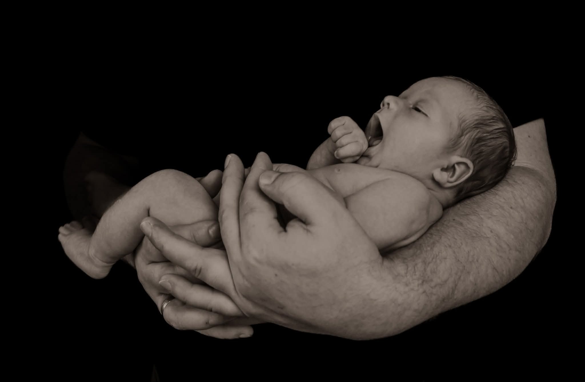 Sepiowe zdjęcie noworodka z otwartymi ustami, trzymanego w dłoniach dorosłego, na czarnym tle. Widoczny pierścionek na palcu.