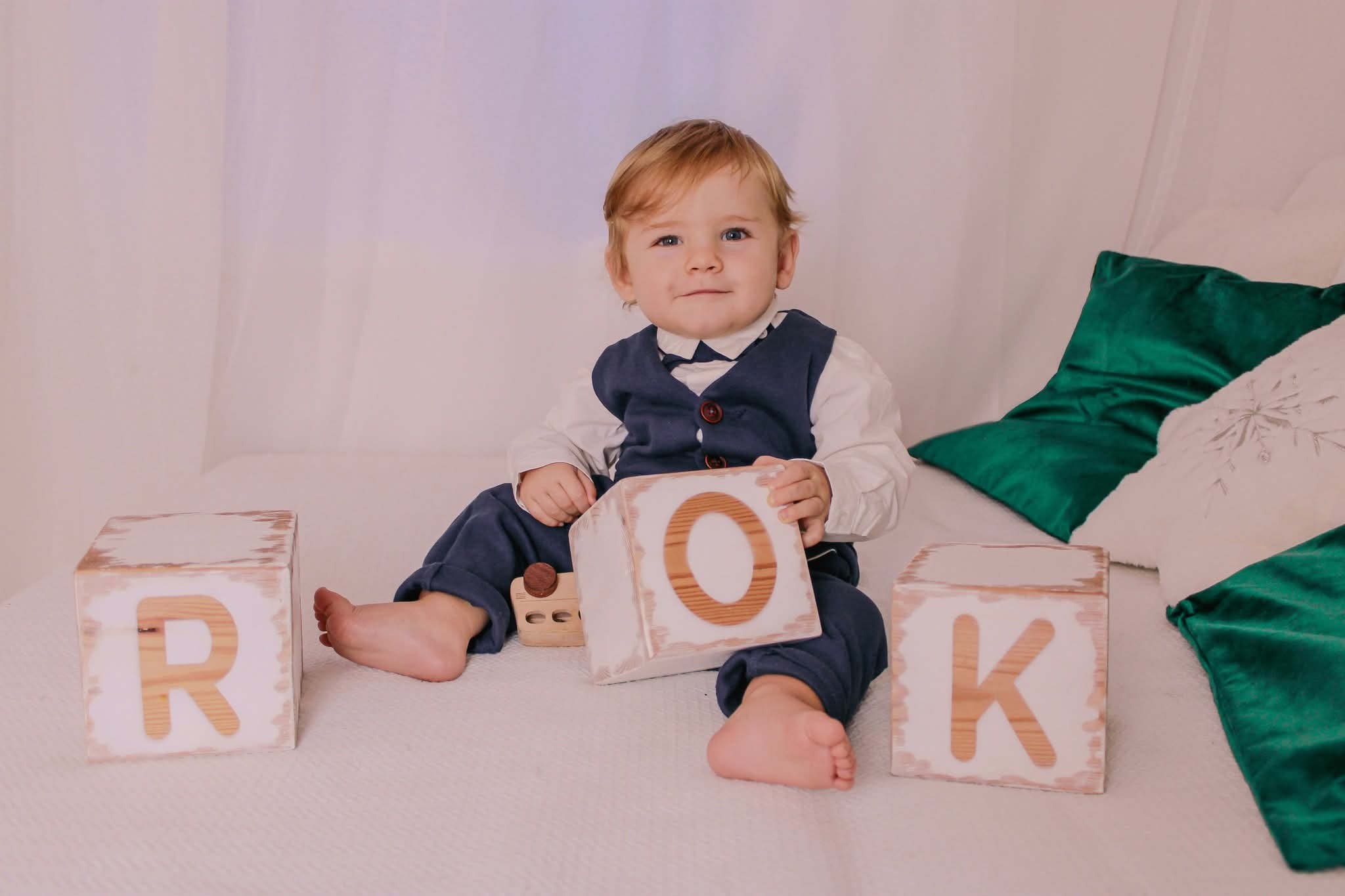 Uroczy chłopiec w eleganckim stroju siedzi na łóżku, otoczony drewnianymi klockami z literami układającymi się w słowo 'ROK'. Jasne tło, miękkie światło.