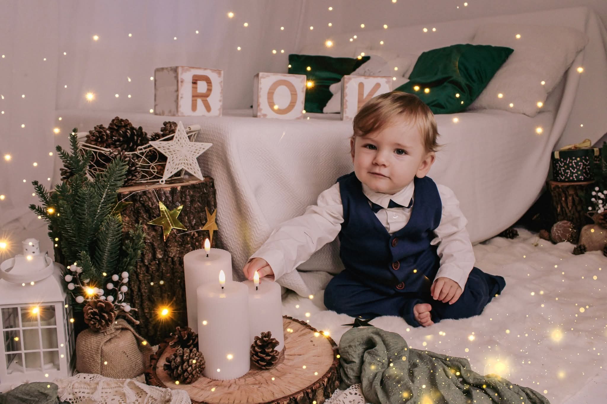 Uroczy chłopiec w granatowym garniturze siedzi na białym futrze, otoczony świecami, dekoracjami świątecznymi i ciepłym, rozproszonym światłem. Sesja noworodkowa.