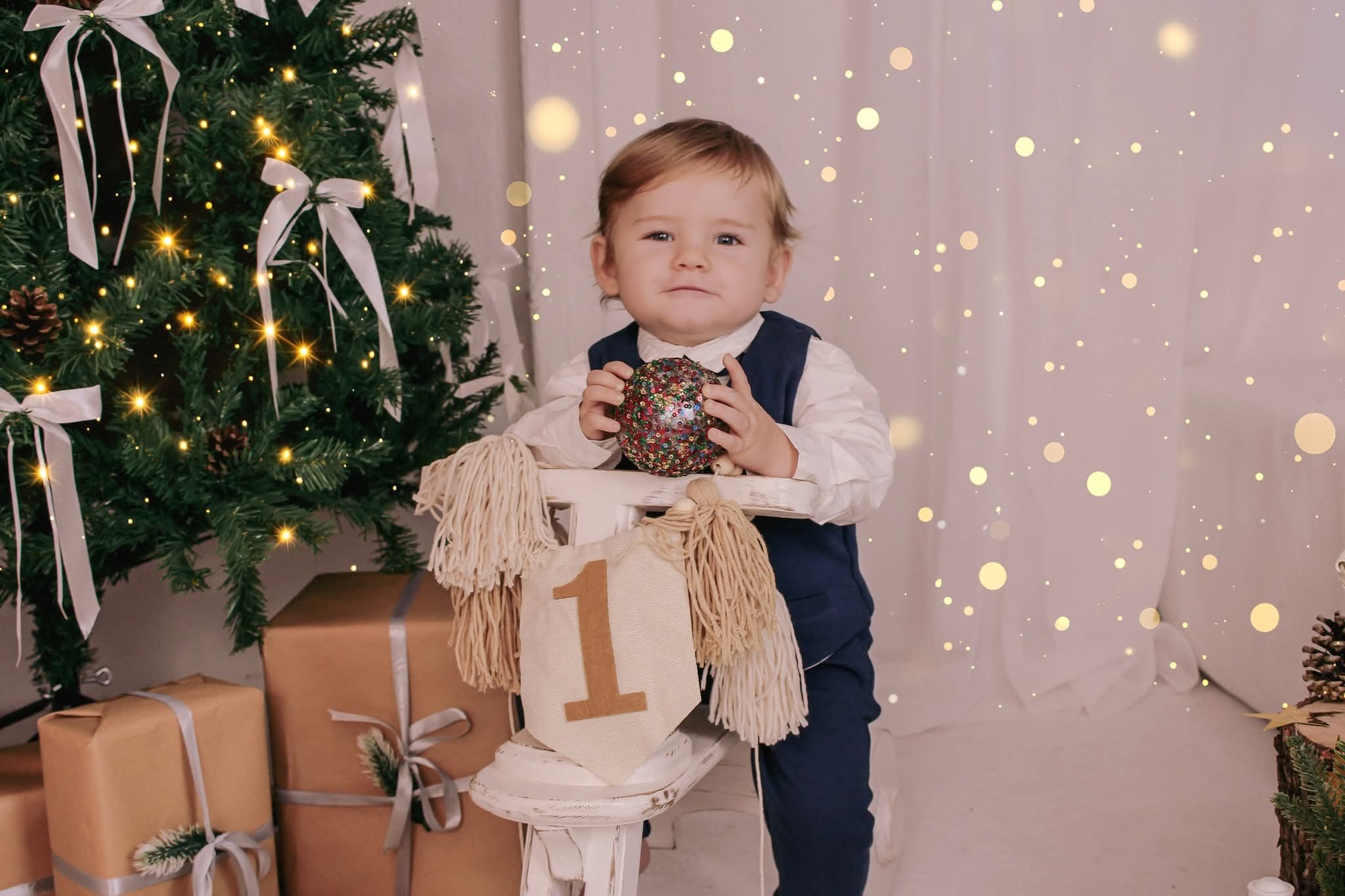 Roczne dziecko w stroju galowym trzyma błyszczącą bombkę na tle choinki, prezentów i dekoracji urodzinowych. Urocza sesja w ciepłych barwach z efektem bokeh.