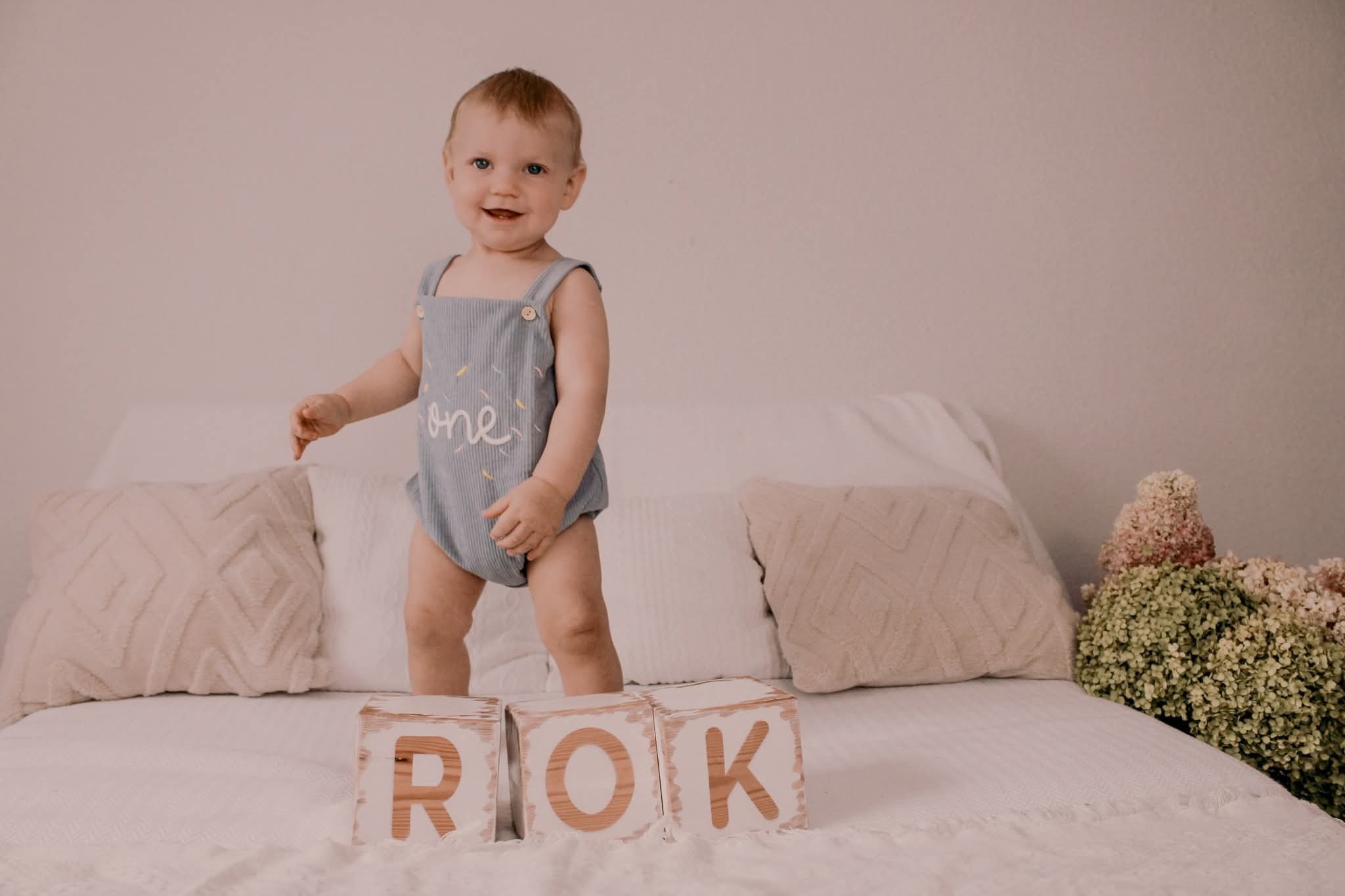 Radosny roczny chłopiec w niebieskim stroju z napisem 'one' stoi na łóżku obok drewnianych klocków z napisem 'ROK', kwiaty w tle, ciepłe oświetlenie.