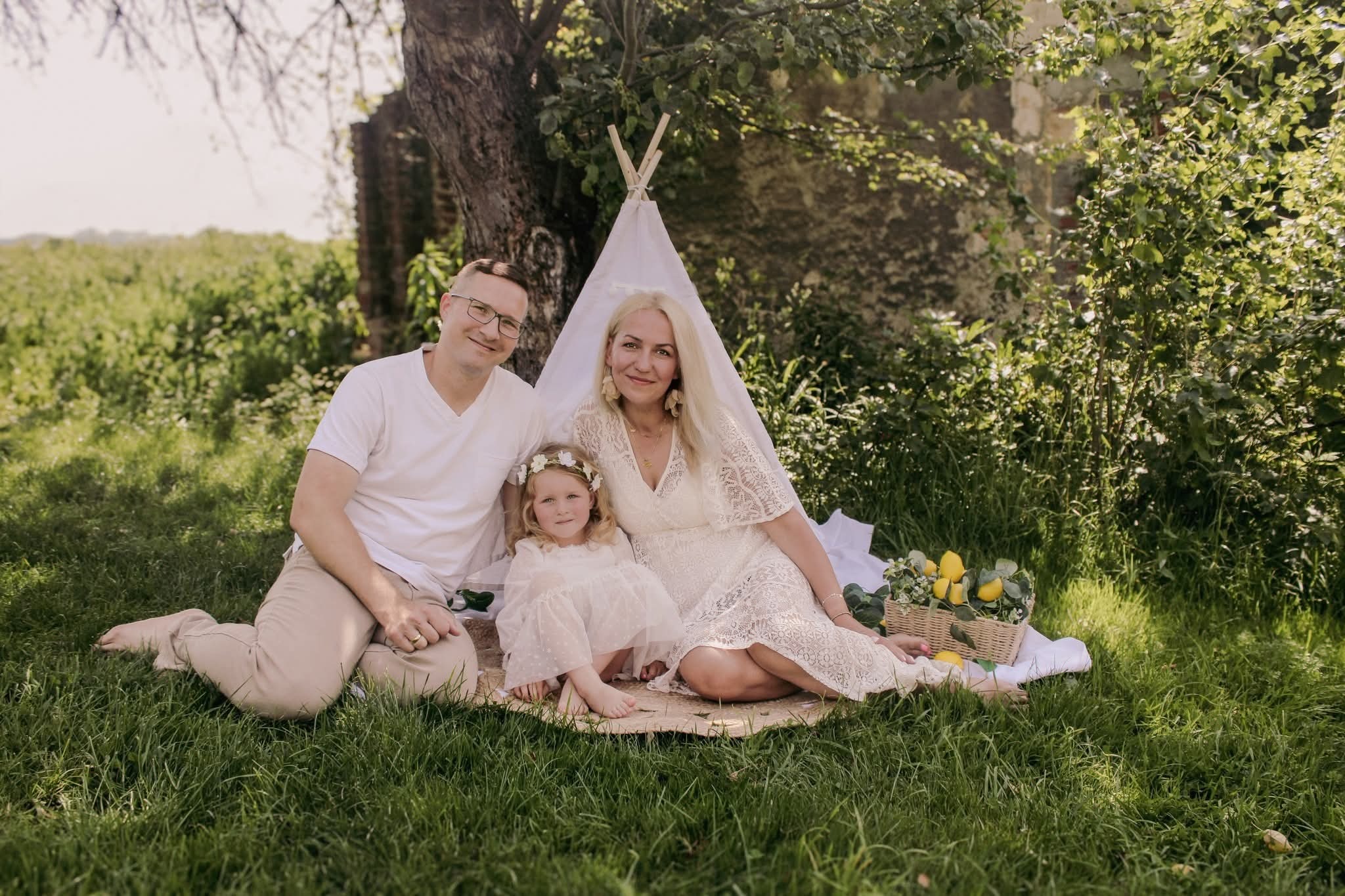 Uśmiechnięta rodzina (mama, tata i córka) w białych ubraniach siedzi na trawie obok małego tipi. W tle drzewa i ruiny. Sielankowa sesja rodzinna w plenerze.