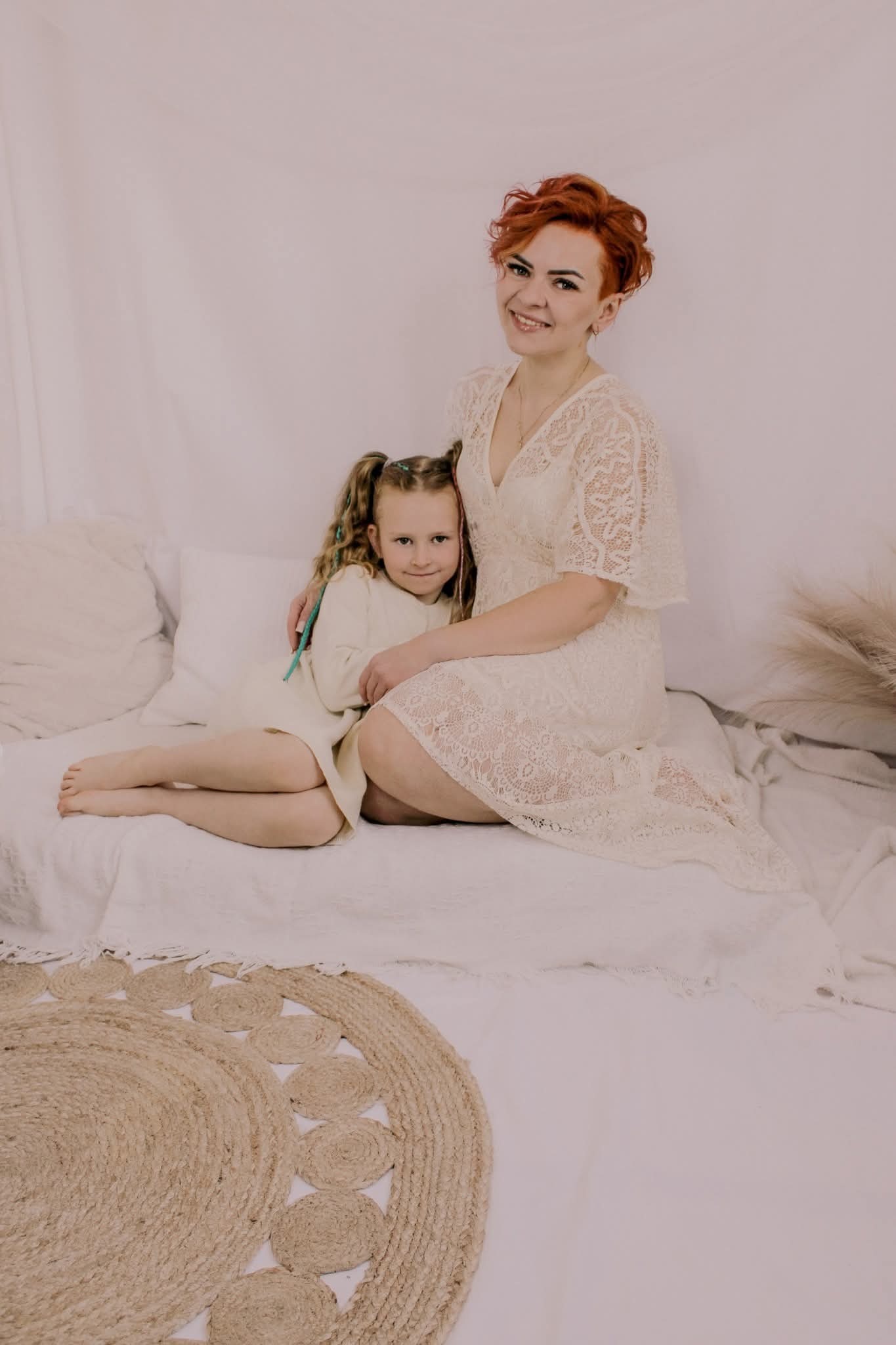 Uśmiechnięta mama z rudymi włosami w koronkowej sukience obejmuje córkę z blond warkoczami na łóżku z jutowym dywanem. Jasne, naturalne światło, neutralne tło.