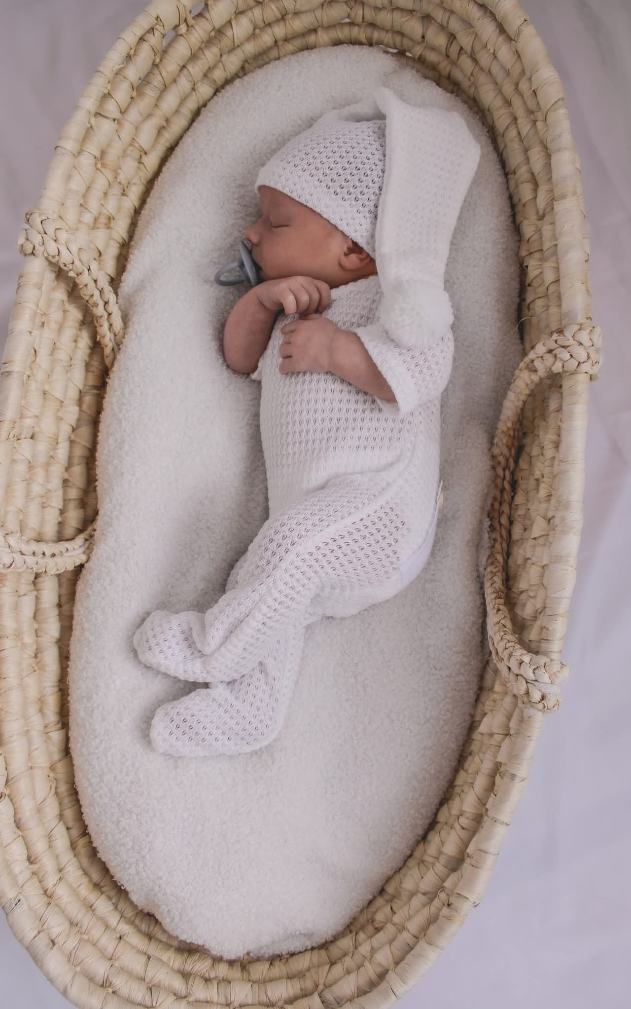 Śpiący noworodek w białym kombinezonie i czapeczce leży w wiklinowym koszu z miękkim, białym wyściełaniem, z błękitnym smoczkiem w ustach. Delikatne światło.