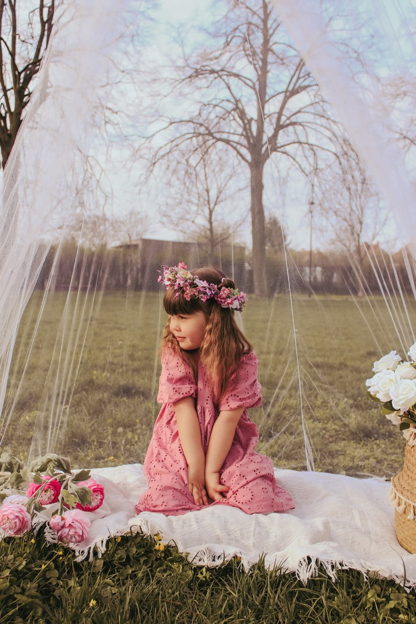 Urocza dziewczynka w różowej sukience i wianku siedzi na kocu w tiulowym namiocie, otoczona kwiatami, zamyślona, w plenerze. Romantyczna sesja w pastelowych barwach.