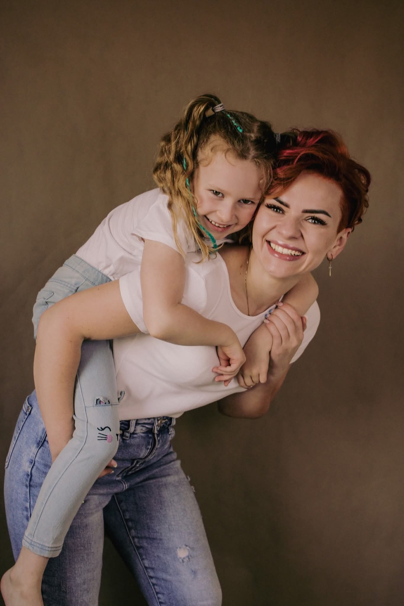 Uśmiechnięta mama z krótkimi, rudymi włosami nosi na plecach córeczkę w jasnych jeansach z aplikacją. Zdjęcie wykonane w Lubaniu na jednolitym, brązowym tle.