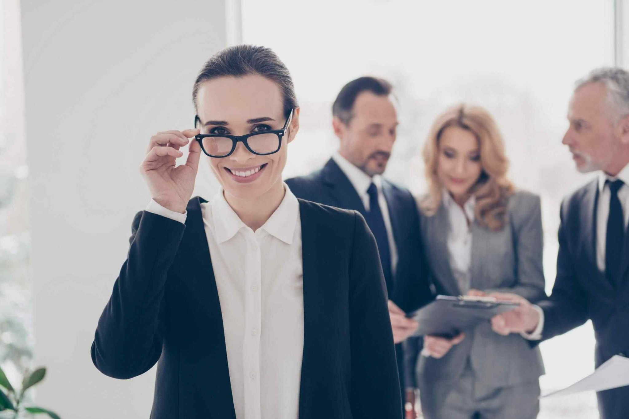 Uśmiechnięta kobieta w okularach i marynarce na pierwszym planie, w tle grupa osób analizująca dokumenty. Jasne, neutralne tło.