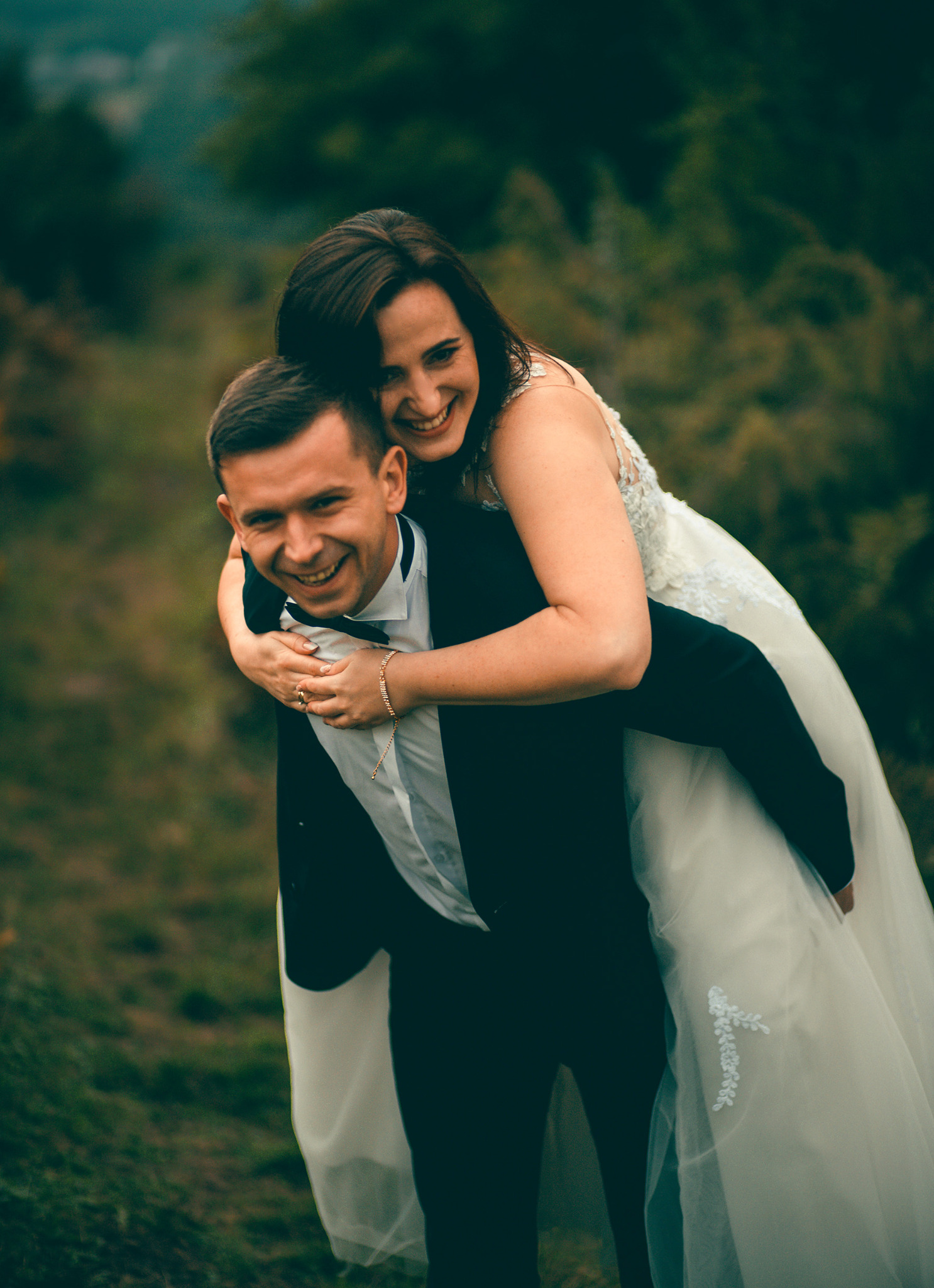 Uśmiechnięta panna młoda na barana u pana młodego w czarnym smokingu, na tle zieleni. Radosna atmosfera sesji ślubnej w plenerze.