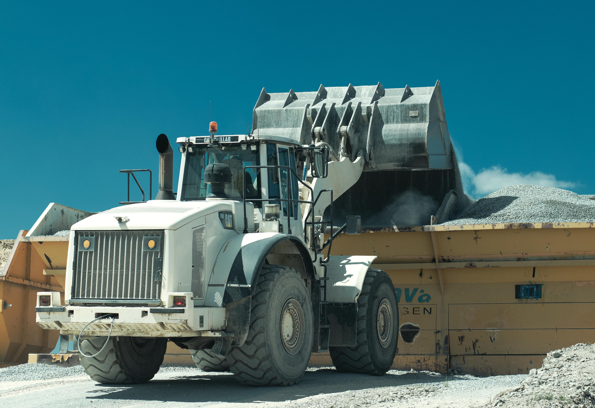Biała ładowarka Caterpillar wysypuje żwir do żółtej naczepy pod błękitnym niebem. Ujęcie z bliska, podkreślające detale maszyny i fakturę materiałów.