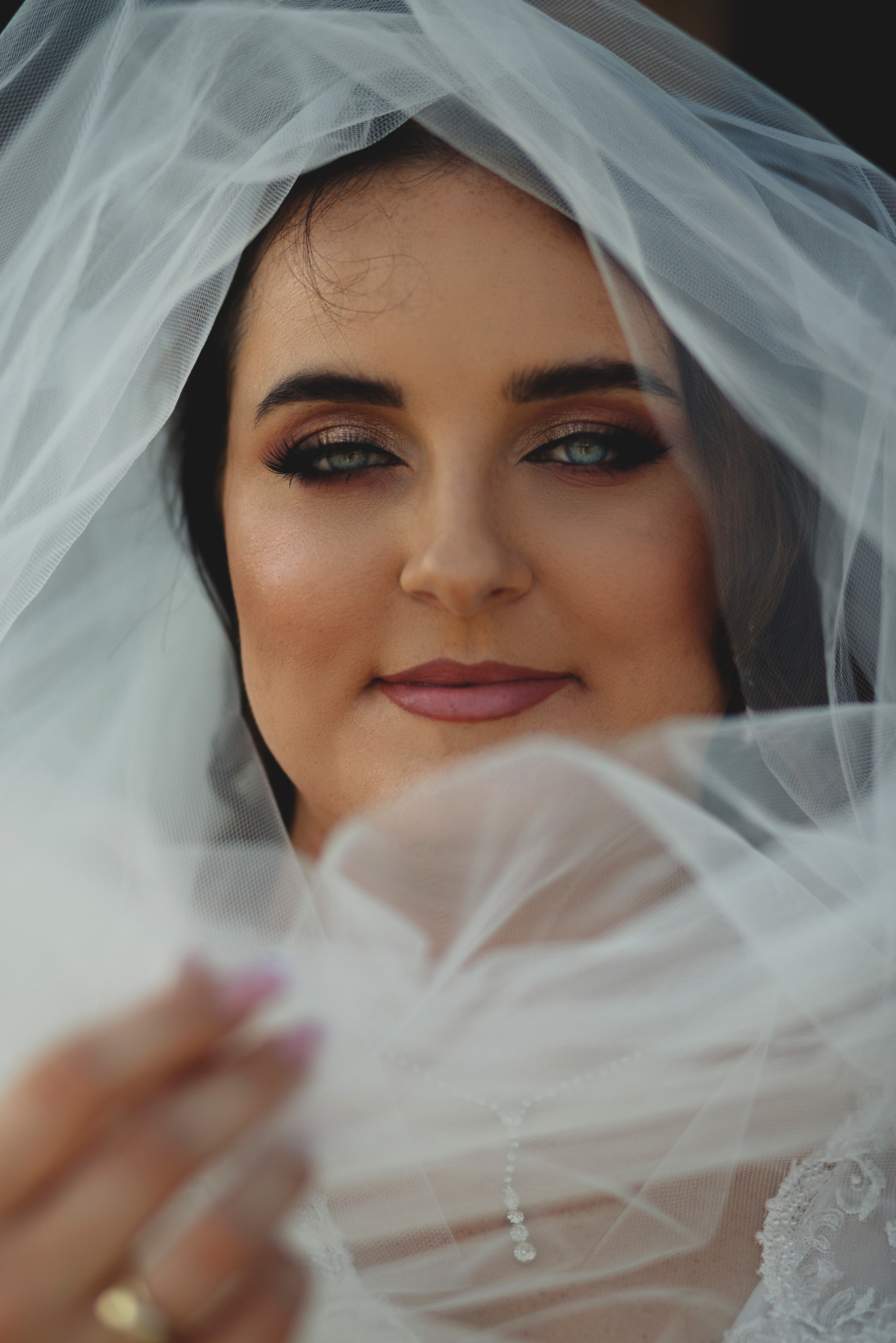 Portret panny młodej z delikatnym makijażem i welonem, z bliska, z widocznym pierścionkiem zaręczynowym i fragmentem sukni ślubnej.