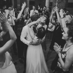 Wyczucie Studio - Czarno-białe zdjęcie pary młodej w tańcu, otoczonej przez gości weselnych klaszczących w dłonie. Para w centrum kadru, z widocznymi detalami sukni ślubnej i garnituru.