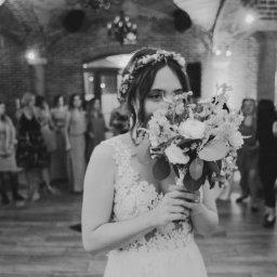 Wyczucie Studio - Czarno-białe zdjęcie panny młodej z bukietem zasłaniającym twarz, w tle goście weselni w ceglanym wnętrzu. Delikatna koronka sukni i wianek na głowie.