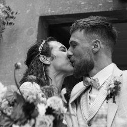 Wyczucie Studio - Czarno-białe zdjęcie całującej się pary młodej na tle kamiennej ściany z kwiatami. Panna młoda w ozdobnej opasce, pan młody w garniturze w kratę i muszce.