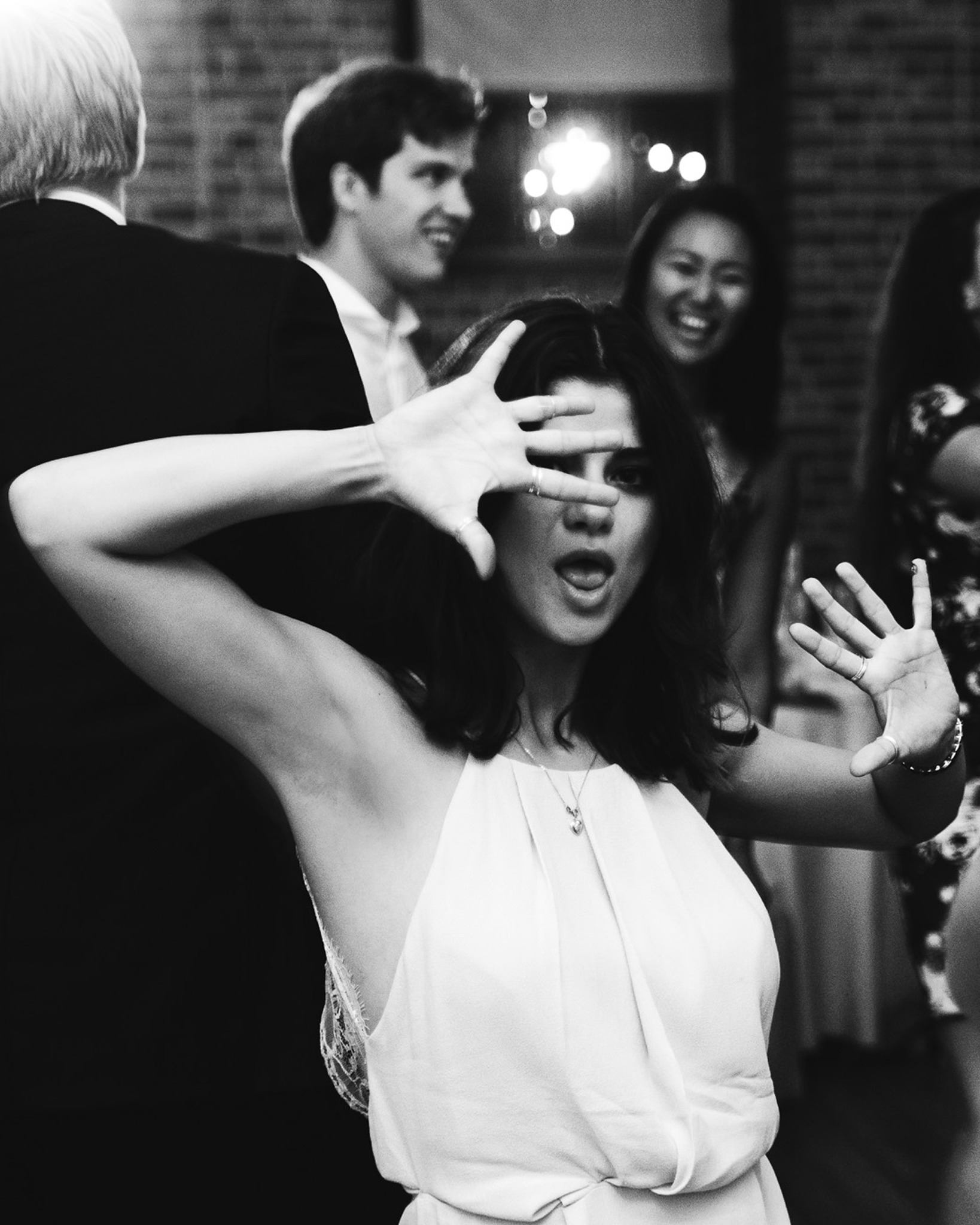 Czarno-białe zdjęcie kobiety z otwartymi ustami, zasłaniającej twarz dłonią na tle rozmytych postaci i świateł, uchwycone podczas wydarzenia.