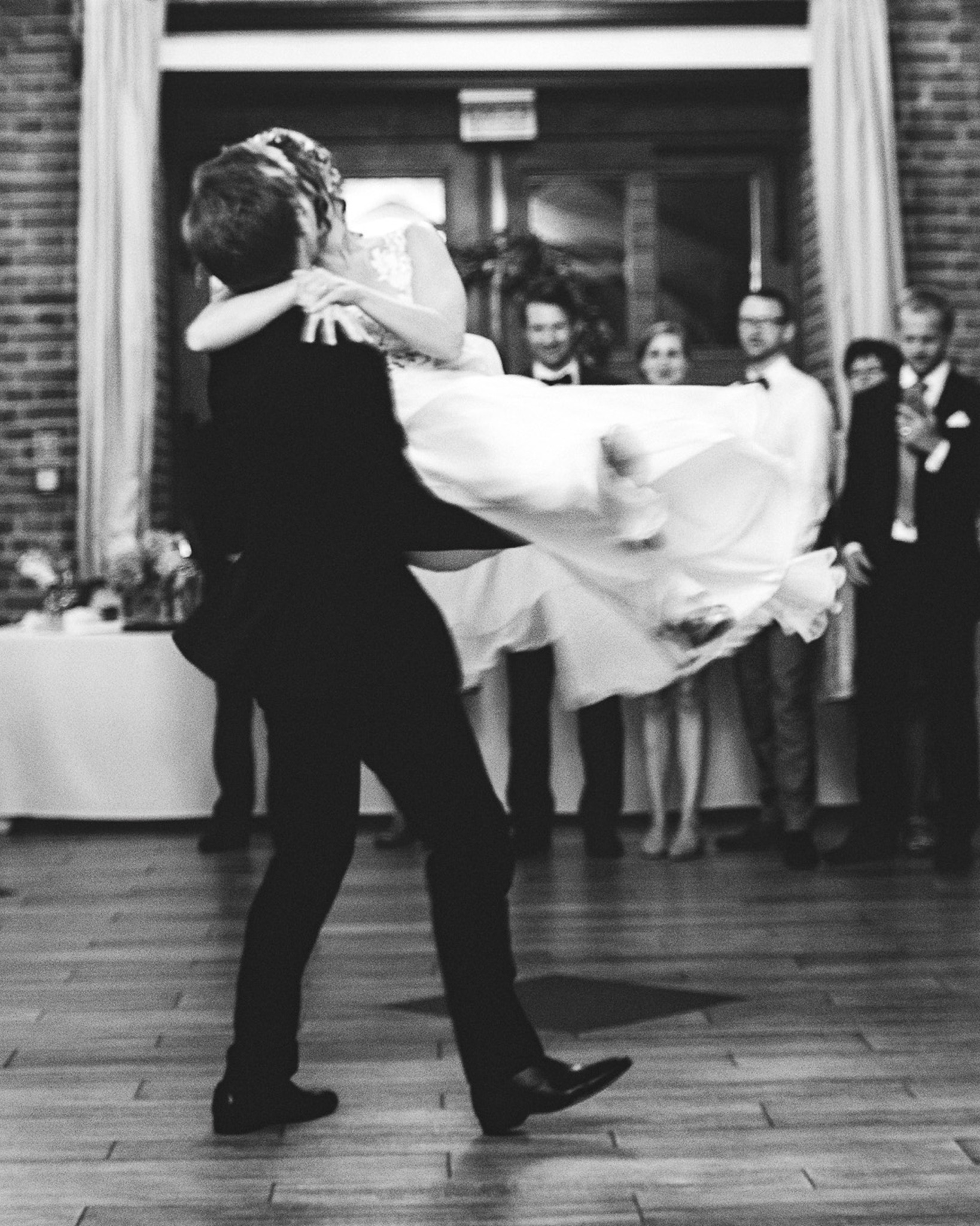 Czarno-białe zdjęcie: Pan młody trzyma pannę młodą w ramionach podczas pierwszego tańca, w tle goście weselni i ceglana ściana.
