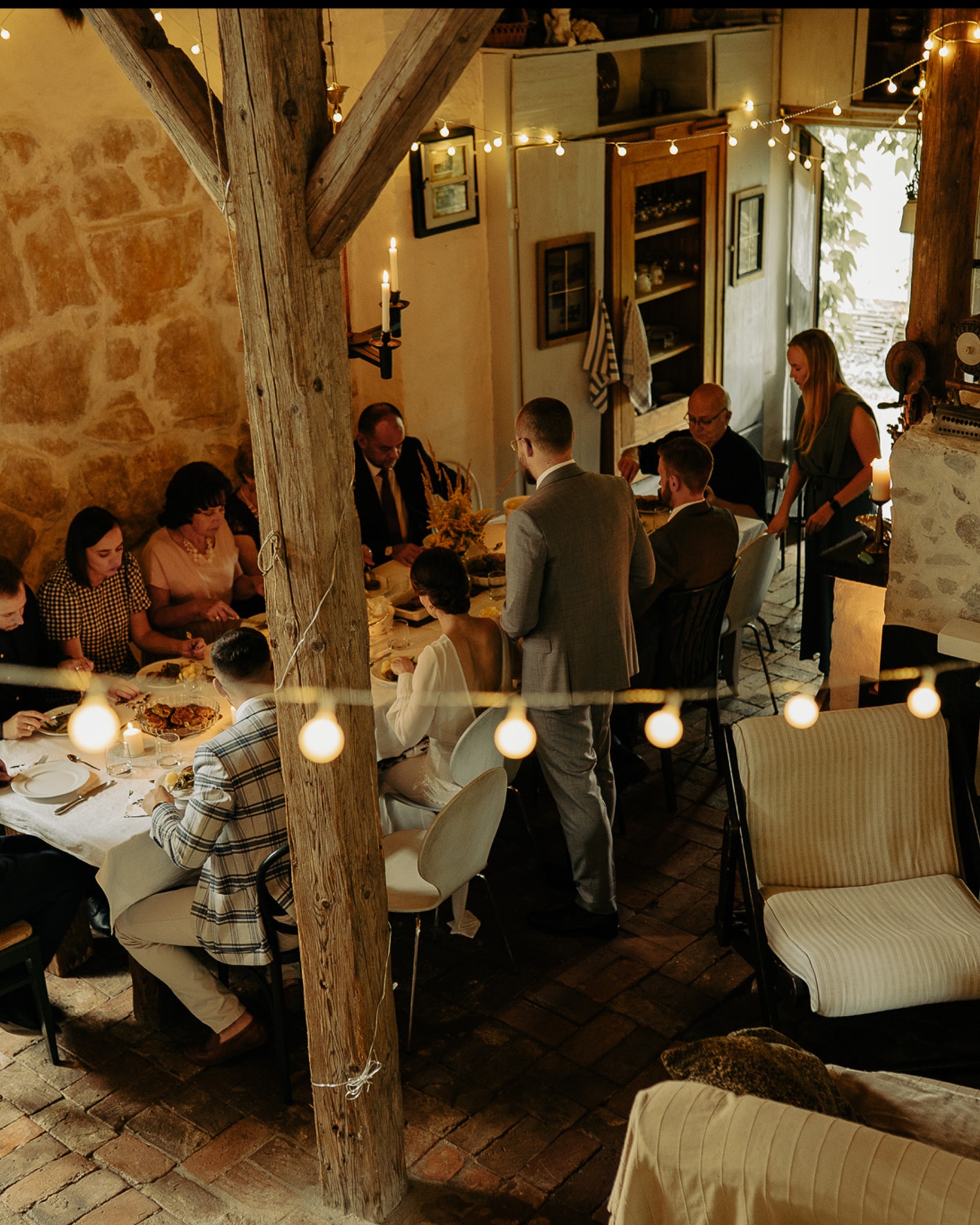 Uroczysty obiad weselny w rustykalnej sali z kamiennymi ścianami, drewnianą konstrukcją i dekoracyjnymi lampkami. Goście siedzą przy stole, w tle bufet i kobieta.