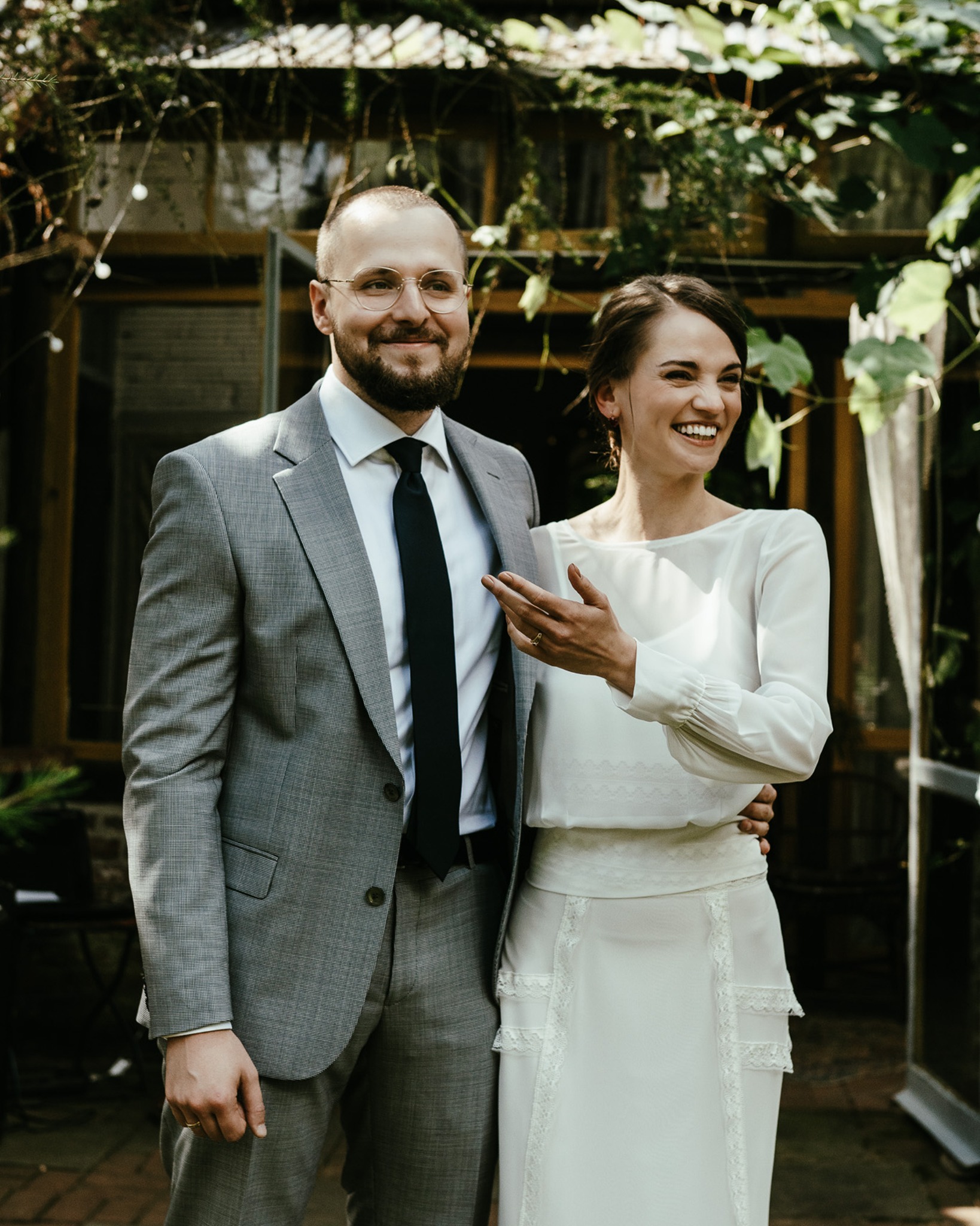 Uśmiechnięta para młoda w plenerze: on w szarym garniturze, ona w białej sukience. Naturalne światło, zielone pnącza w tle, ciepła atmosfera.