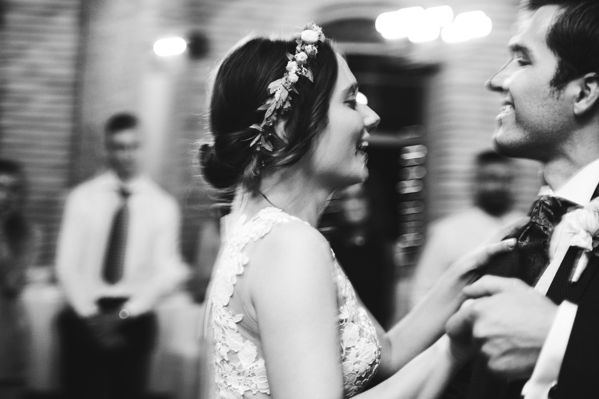 Czarno-białe zdjęcie uśmiechniętej panny młodej w koronie z kwiatów, tańczącej z panem młodym w garniturze. Rozmyte tło z gośćmi.