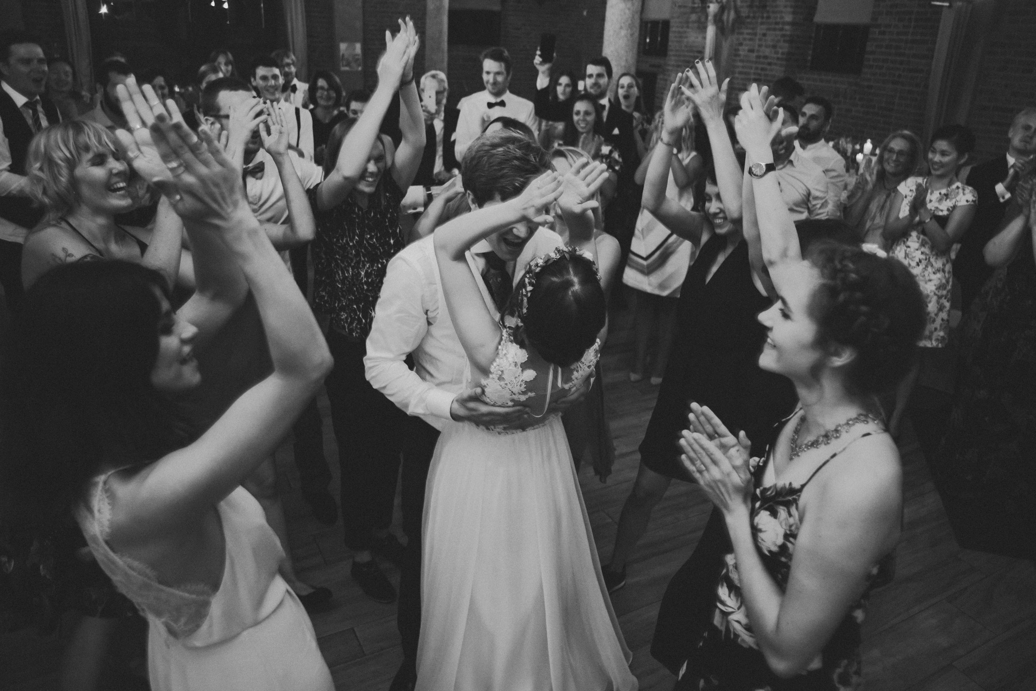 Czarno-białe zdjęcie pary młodej w tańcu, otoczonej przez gości weselnych klaszczących w dłonie. Para w centrum kadru, z widocznymi detalami sukni ślubnej i garnituru.
