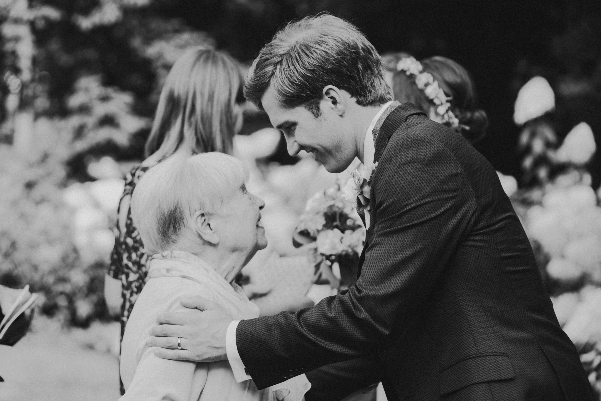 Czarno-białe zdjęcie pana młodego w ciemnym garniturze, który z czułością obejmuje starszą kobietę podczas uroczystości ślubnej, z rozmyciem w tle.
