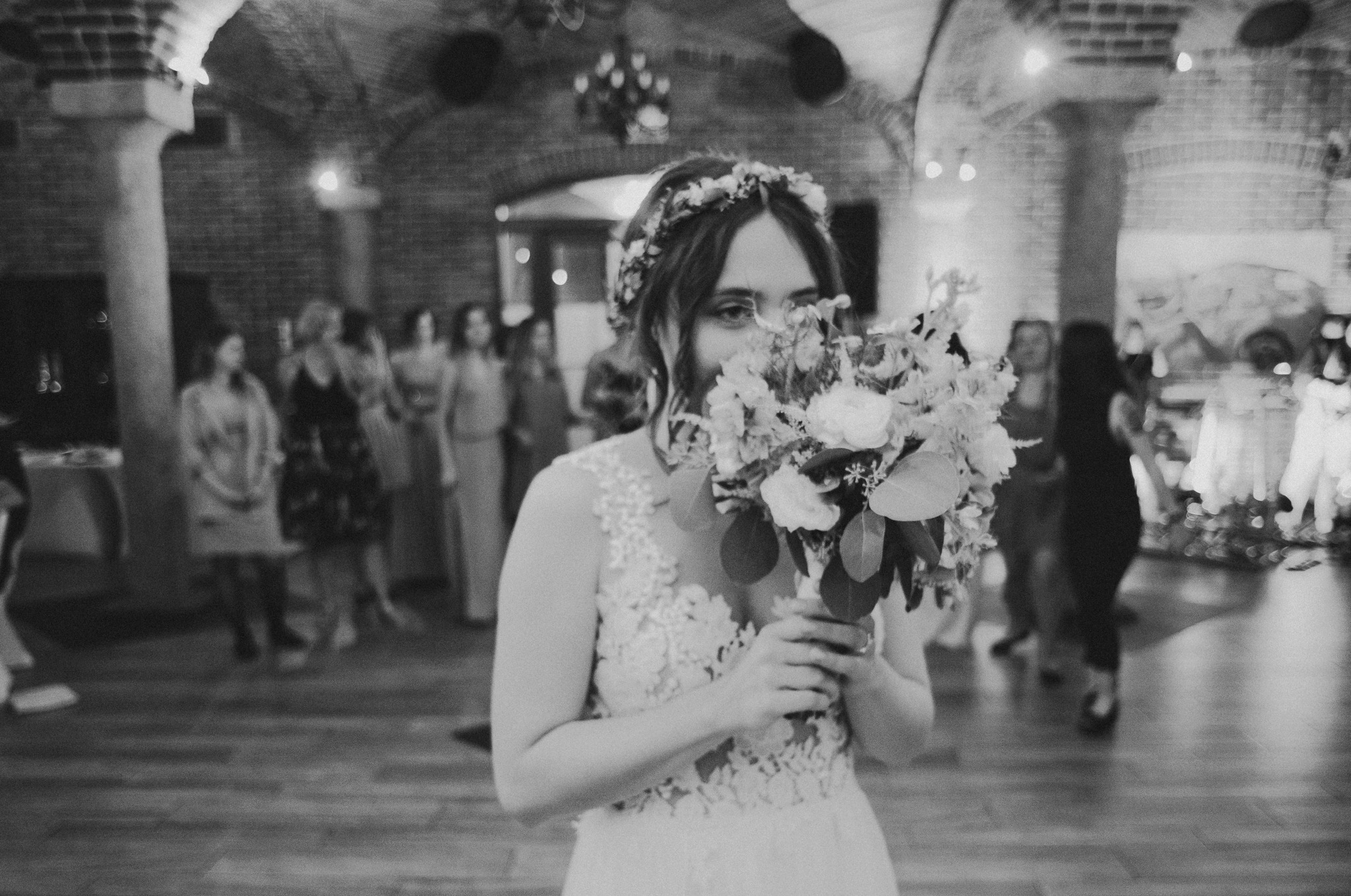 Czarno-białe zdjęcie panny młodej z bukietem zasłaniającym twarz, w tle goście weselni w ceglanym wnętrzu. Delikatna koronka sukni i wianek na głowie.