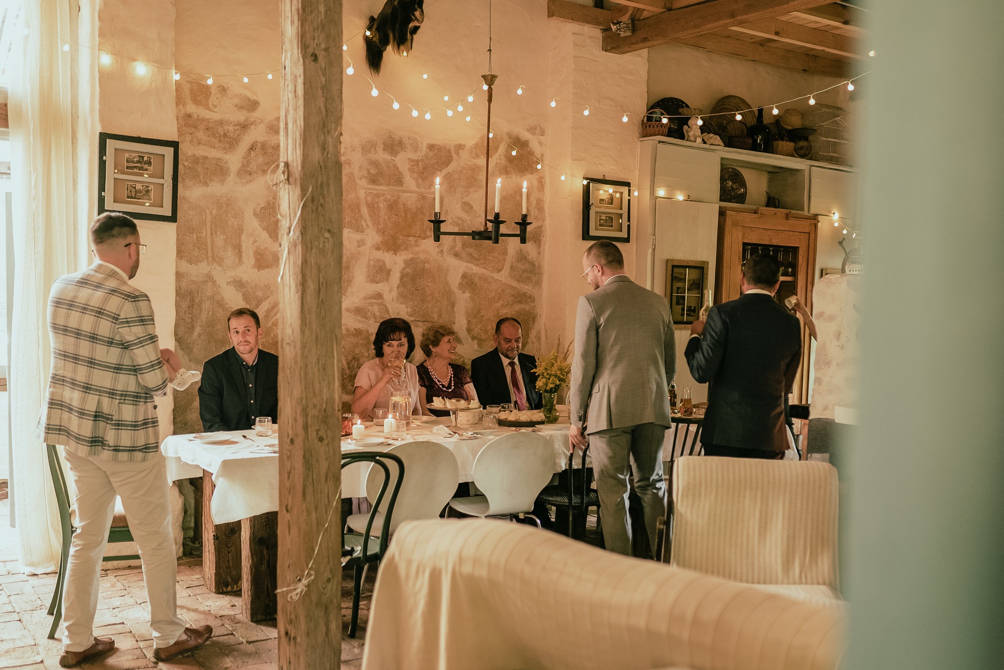 Uroczysty obiad w rustykalnej sali: goście siedzą przy stole, oświetlonym świecami i girlandami, w tle kamienna ściana i dekoracje. Mężczyźni w garniturach w ruchu.
