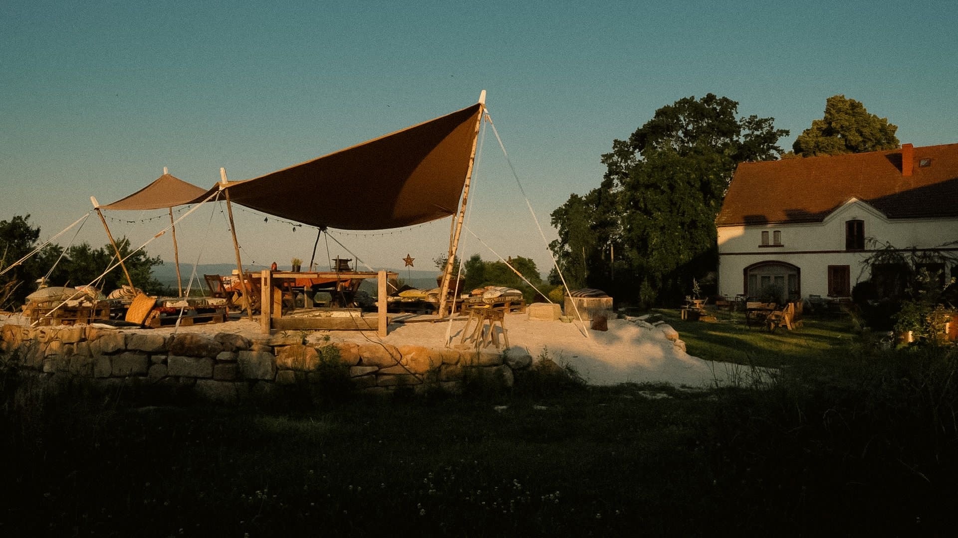 Urokliwa przestrzeń eventowa na zewnątrz z namiotem w stylu boho, drewnianymi meblami i kamiennym murem, w tle biały dom z czerwoną dachówką, oświetlona ciepłym światłem.