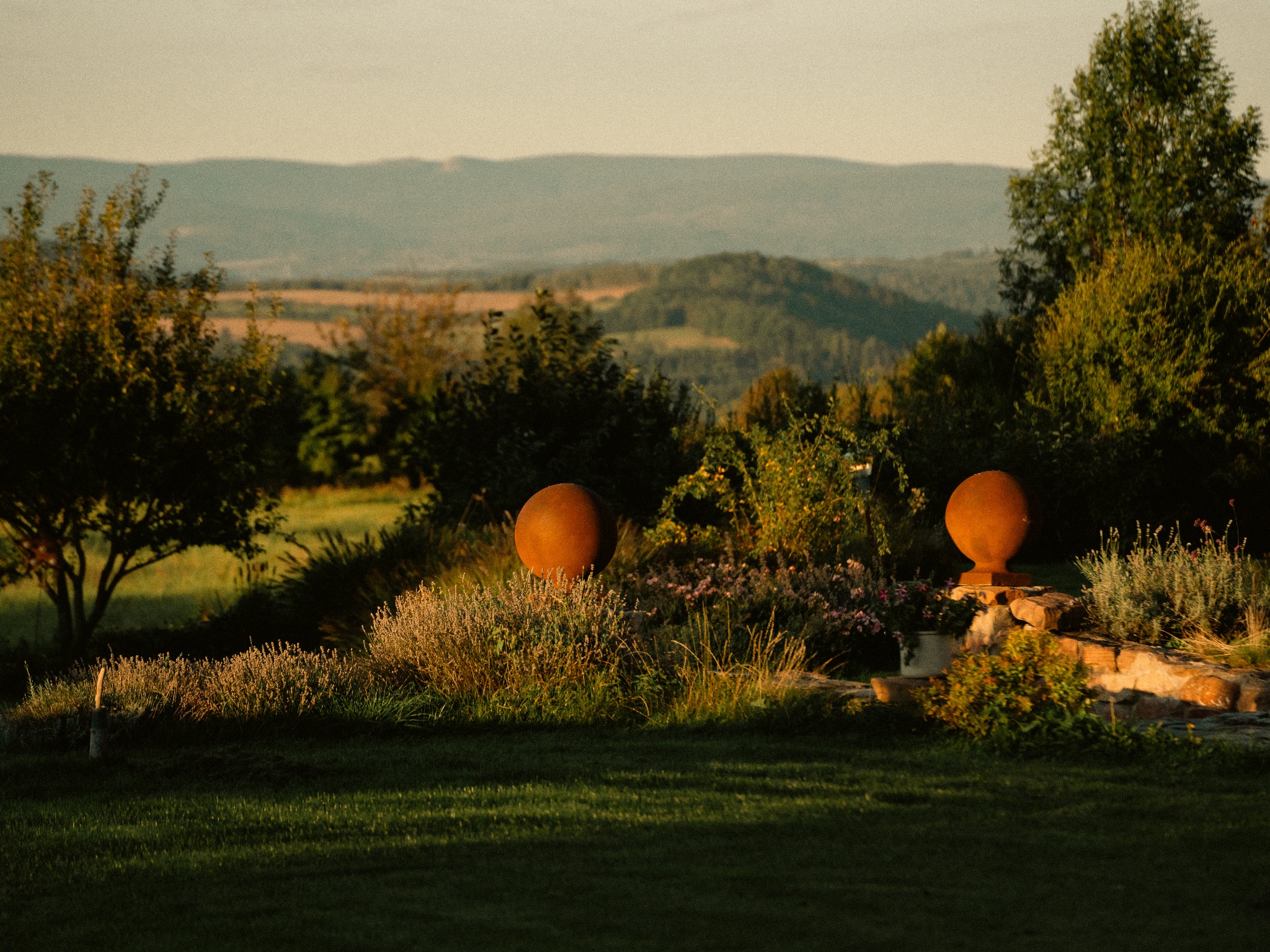 Rustykalny ogród z kulistymi dekoracjami i widokiem na wzgórza w ciepłym, zachodzącym słońcu. Kamienna rabata z lawendą i innymi roślinami.