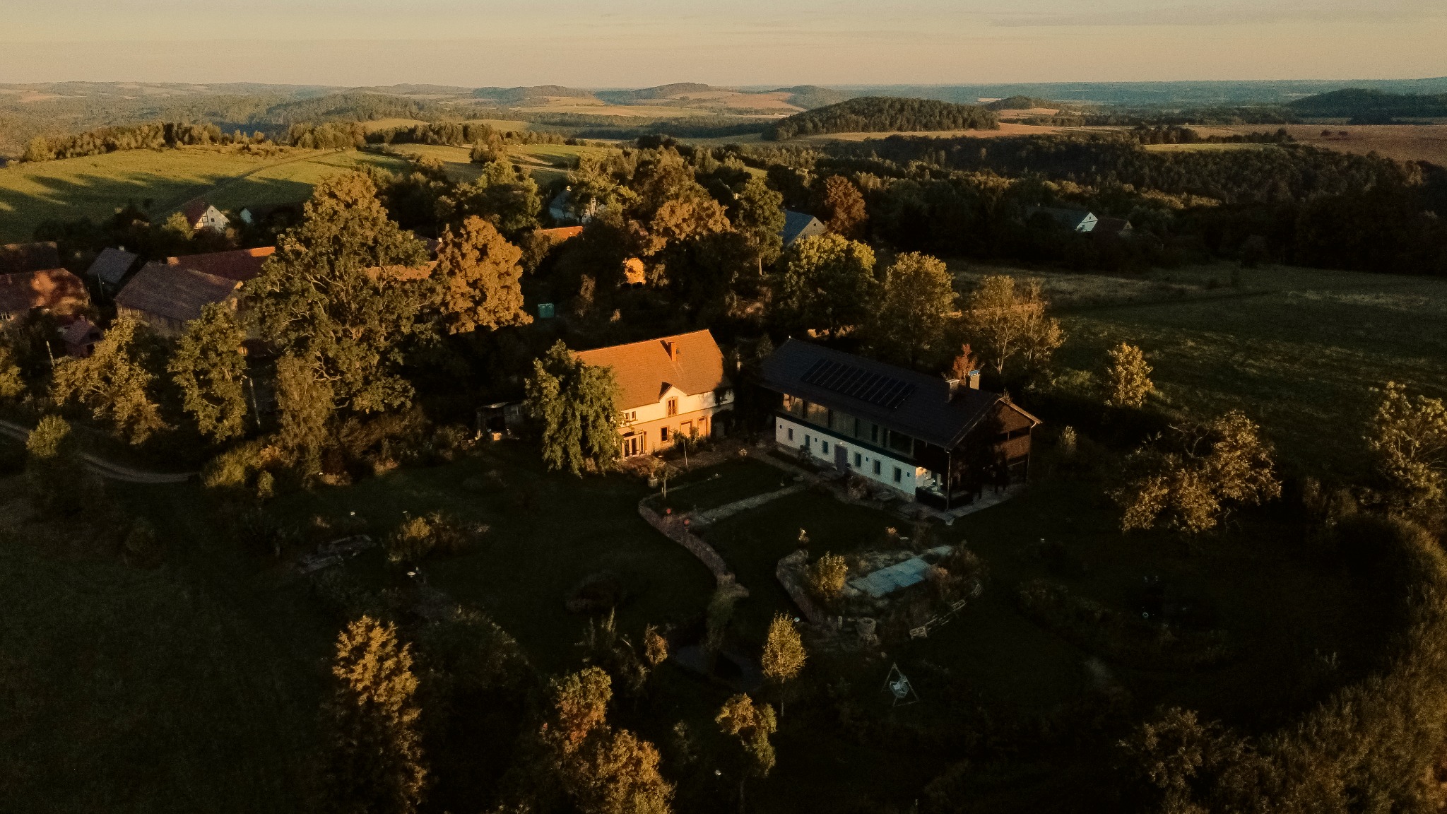 Panoramiczny widok z lotu ptaka na dwa domy jednorodzinne otoczone zielenią, w tle pagórkowaty krajobraz oświetlony ciepłym światłem zachodzącego słońca.