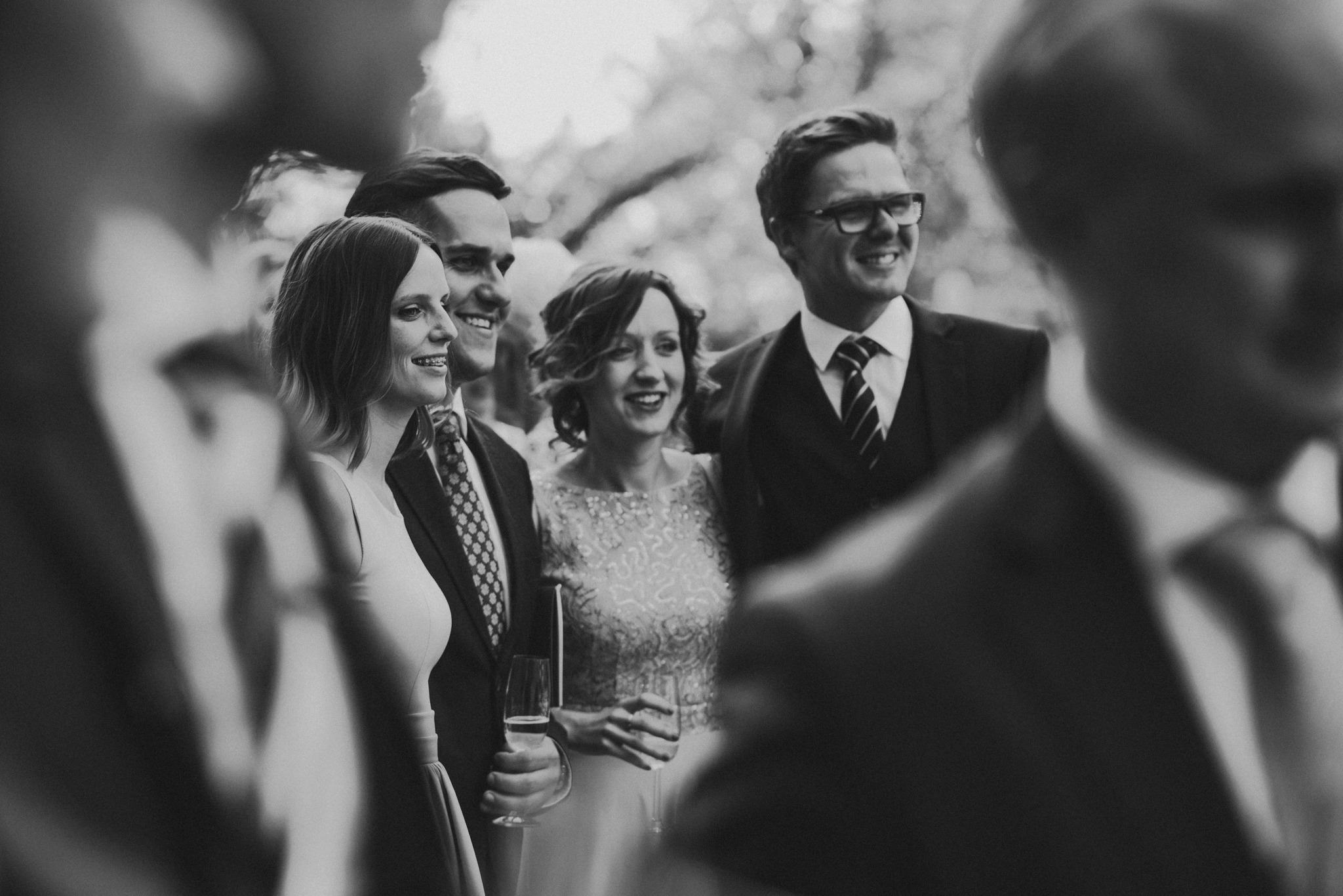Czarno-białe zdjęcie uśmiechniętych gości weselnych, z kieliszkami szampana, w eleganckich strojach, z delikatnym rozmyciem tła, uchwycone w naturalnym świetle.