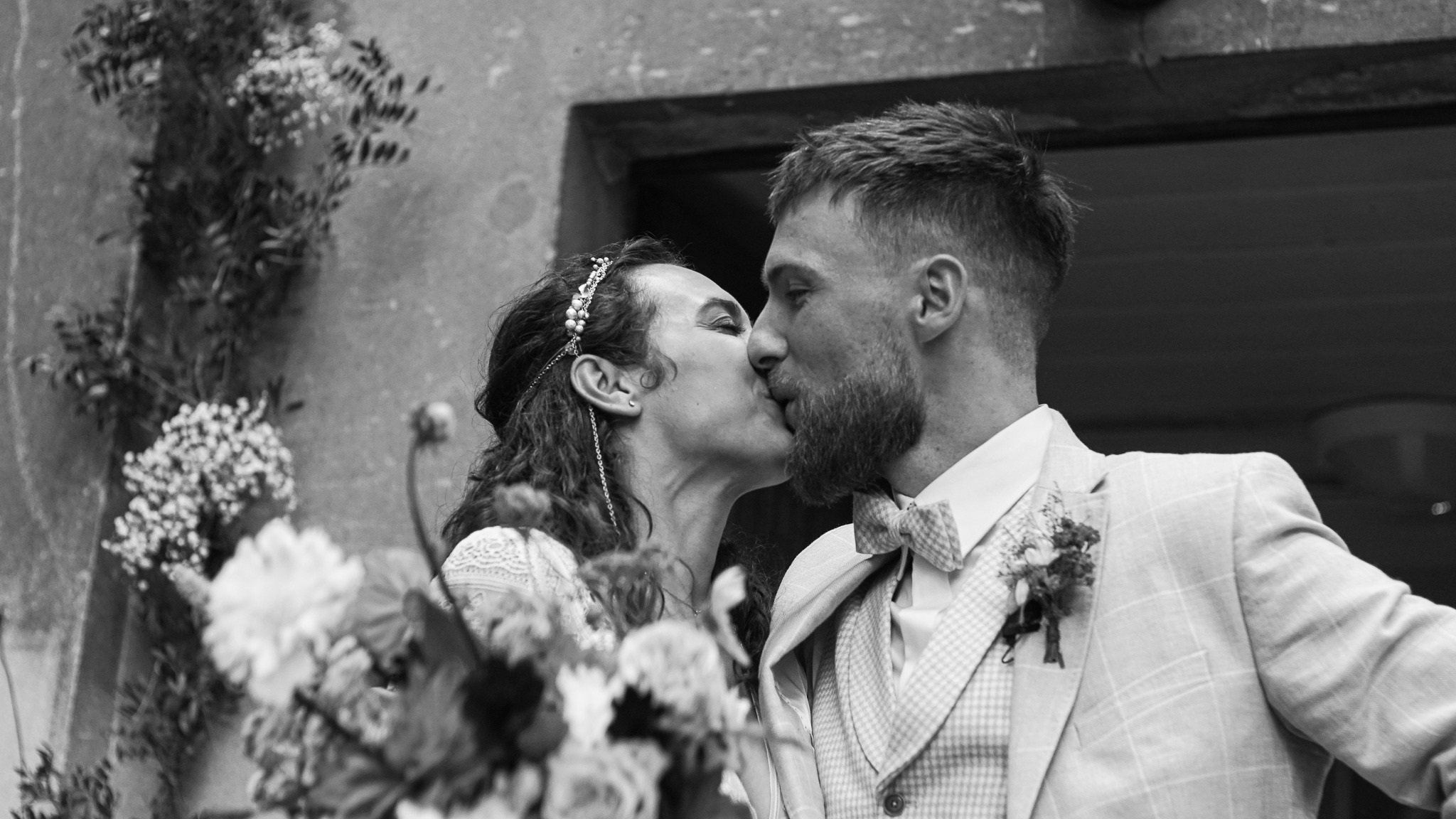 Czarno-białe zdjęcie całującej się pary młodej na tle kamiennej ściany z kwiatami. Panna młoda w ozdobnej opasce, pan młody w garniturze w kratę i muszce.