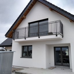 ZAKŁAD USŁUG SPAWALNICZYCH JAROSŁAW BORKOWSKI - Nowoczesny dom z balkonem i czarną, metalową balustradą. Minimalistyczny design, biała elewacja, ciemna dachówka. Widok z dołu, pochmurne niebo w tle.