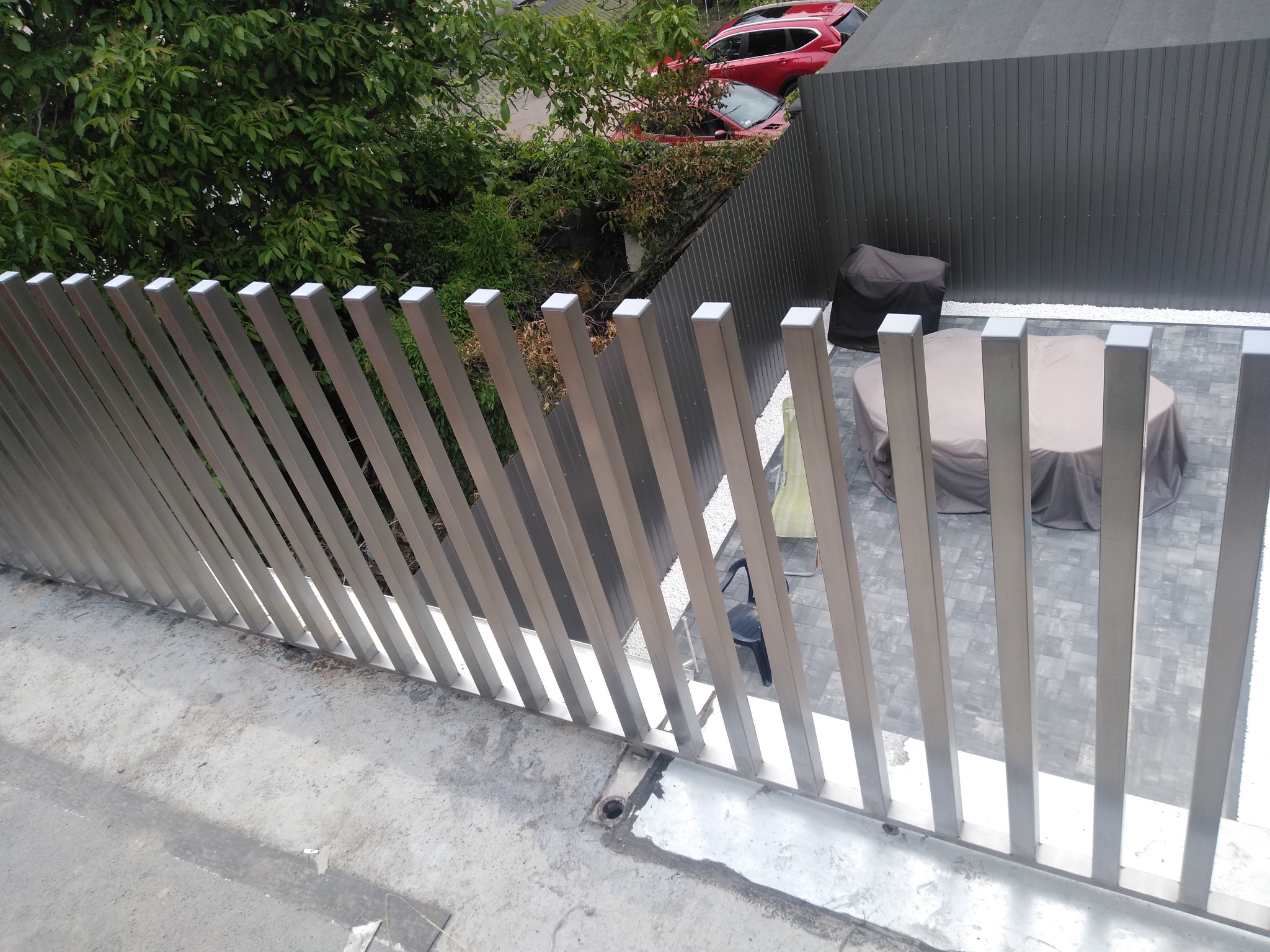 Nowoczesna balustrada balkonowa z metalowych, pionowych profili o kwadratowym przekroju, z widokiem na taras i zieleń. Minimalistyczny design, jasne wykończenie.