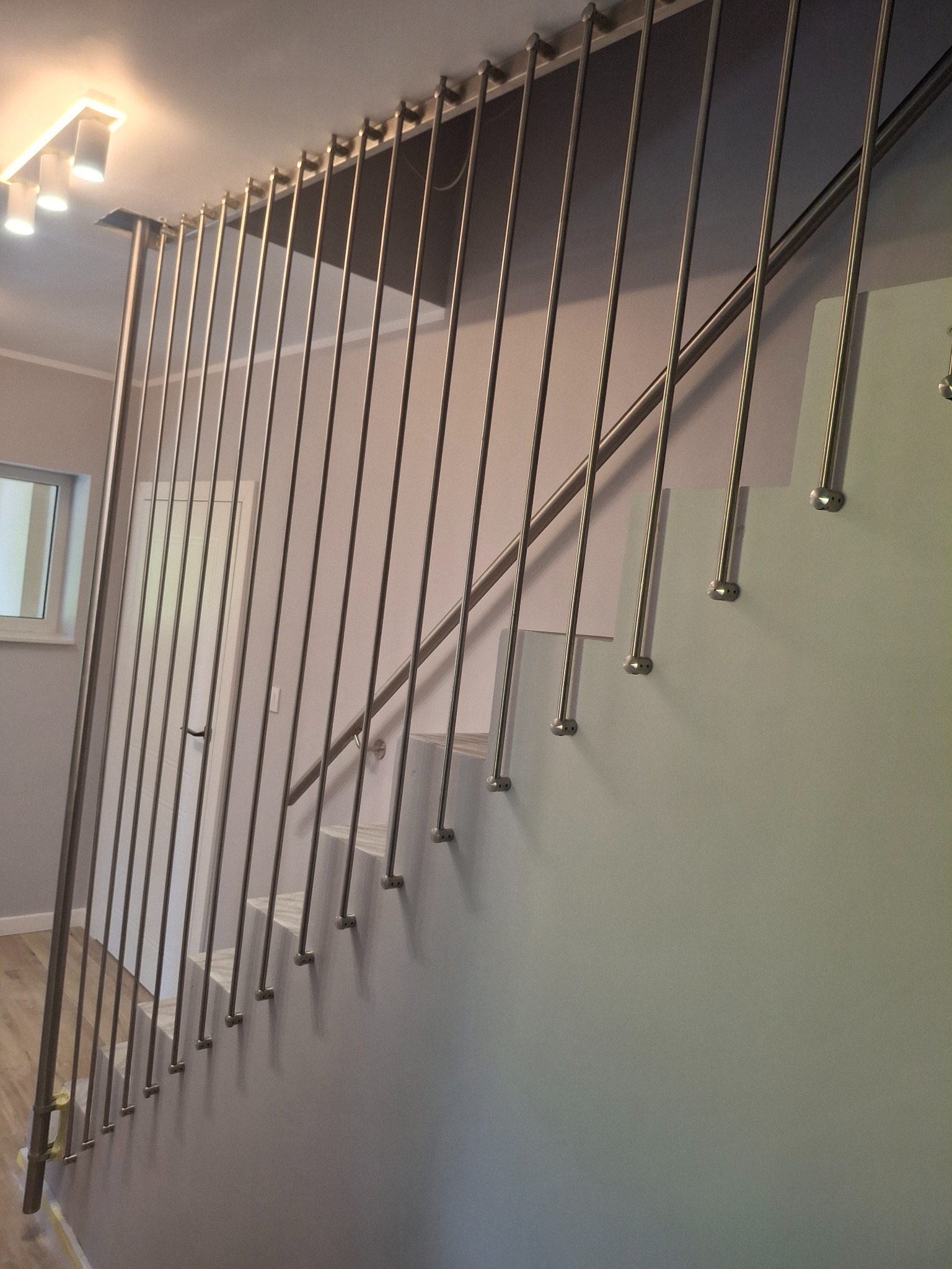 Nowoczesne schody z balustradą ze stali nierdzewnej, pionowe pręty, minimalistyczny design, jasne wnętrze, widok z dołu na górę.