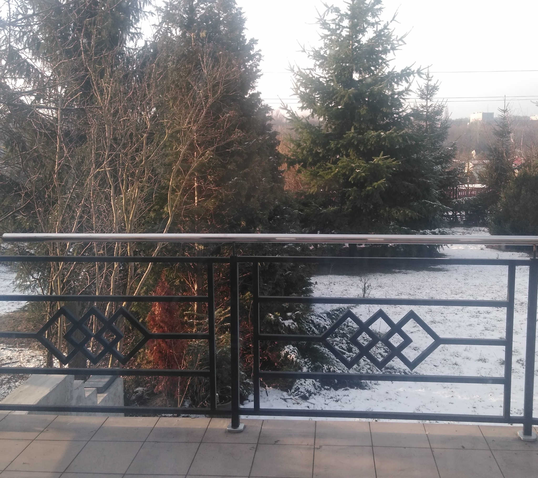 Metalowa balustrada balkonowa z geometrycznym wzorem rombów na tle zimowego krajobrazu z drzewami iglastymi i śniegiem. Widok z balkonu na zewnątrz.