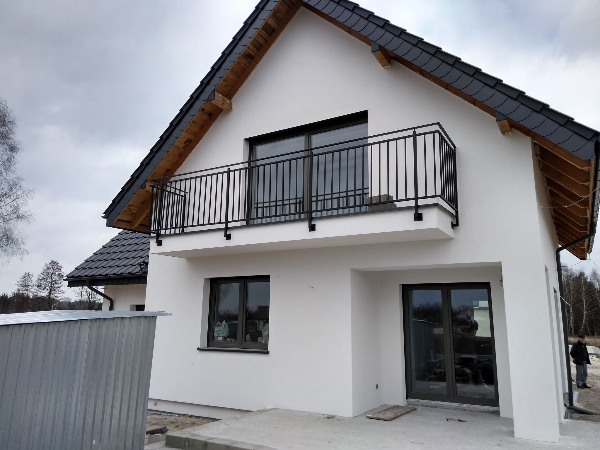 Nowoczesny dom z balkonem i czarną, metalową balustradą. Minimalistyczny design, biała elewacja, ciemna dachówka. Widok z dołu, pochmurne niebo w tle.