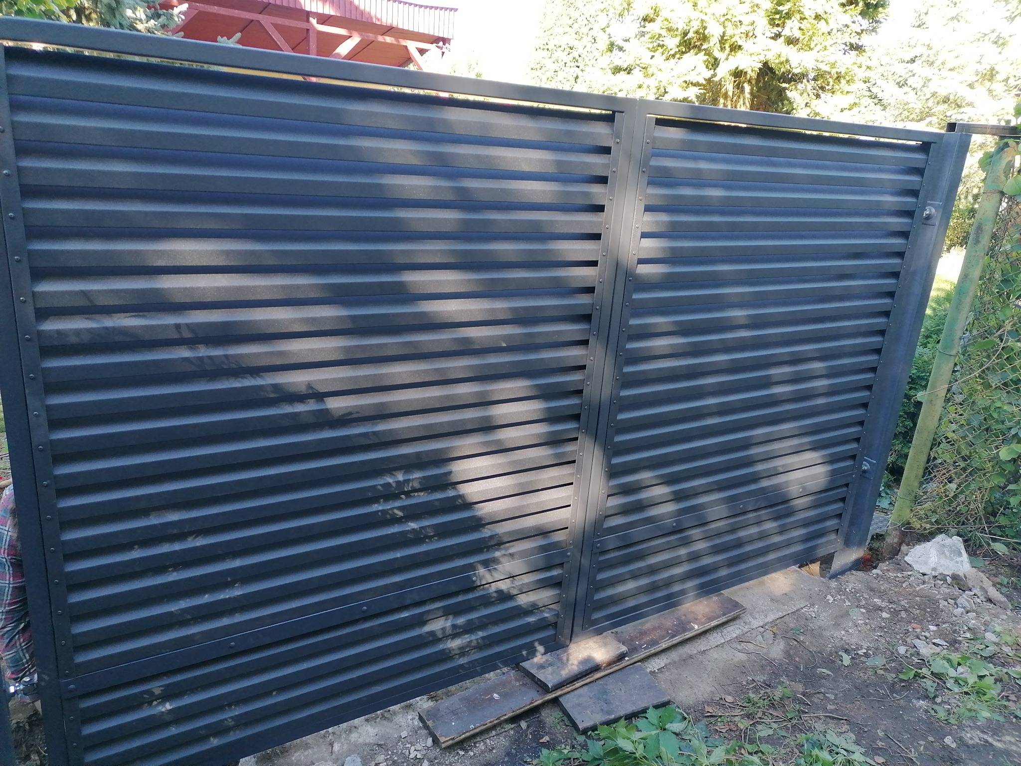 Nowoczesna, pozioma brama wjazdowa z metalowych paneli w kolorze grafitowym, zamontowana na posesji z widoczną zielenią w tle. Brama z bliska, z detalami łączeń.