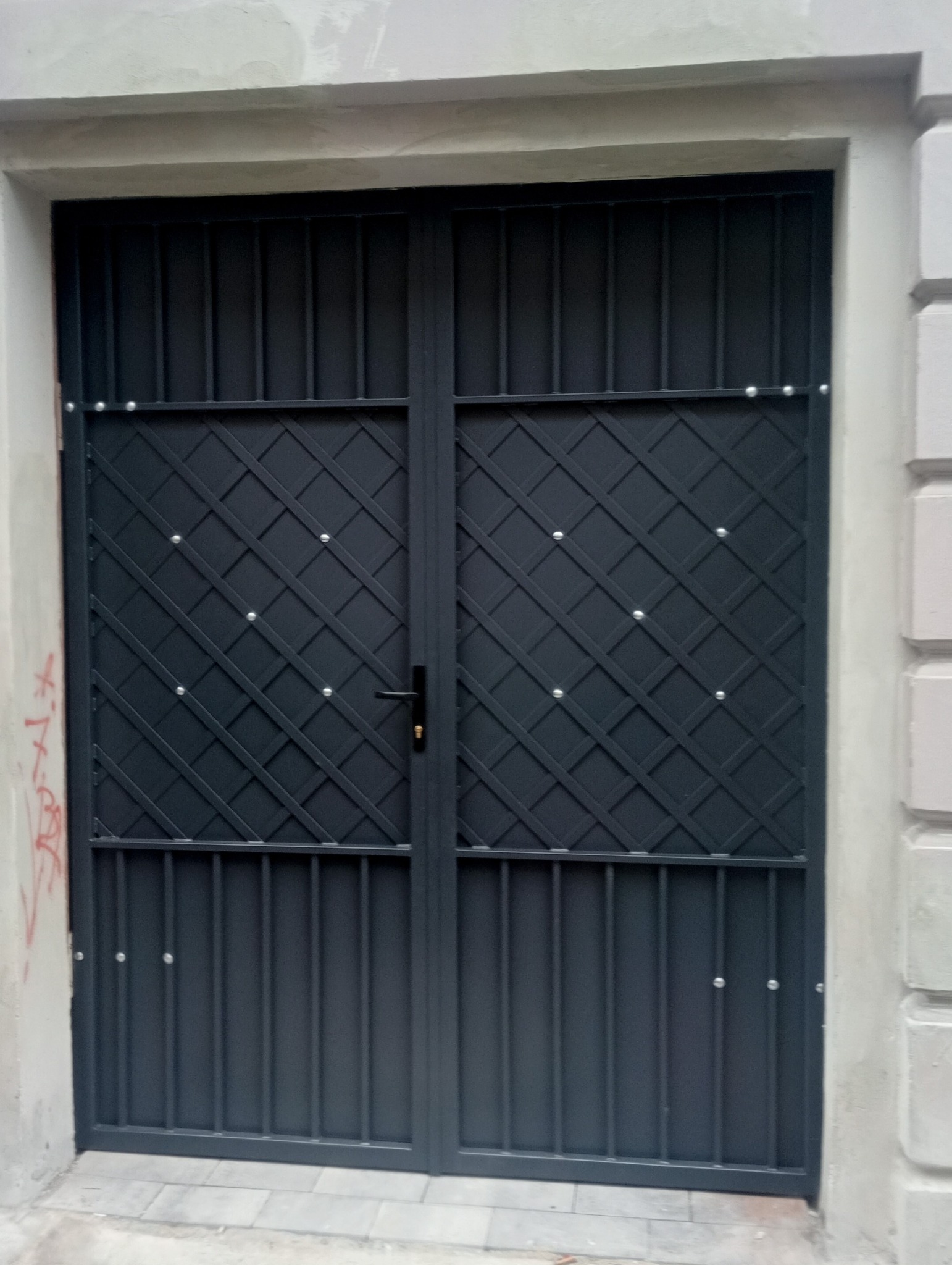 Metalowa brama wjazdowa w kolorze grafitowym, z kratownicą w górnej części i pionowymi prętami w dolnej, zamontowana w jasnym murze.