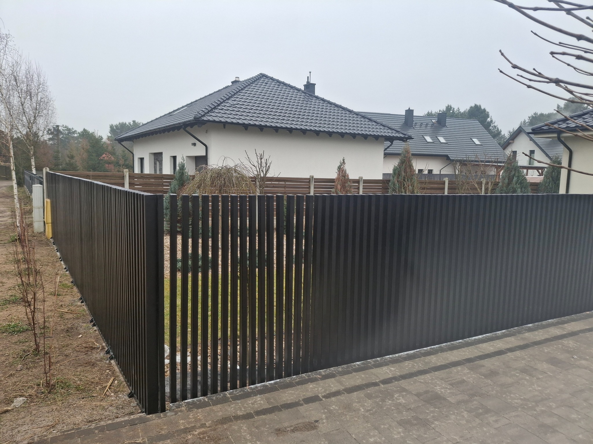 Nowoczesne, pionowe ogrodzenie metalowe w kolorze grafitowym, z widocznymi przerwami między elementami, na tle domu z ciemnym dachem i kostki brukowej.