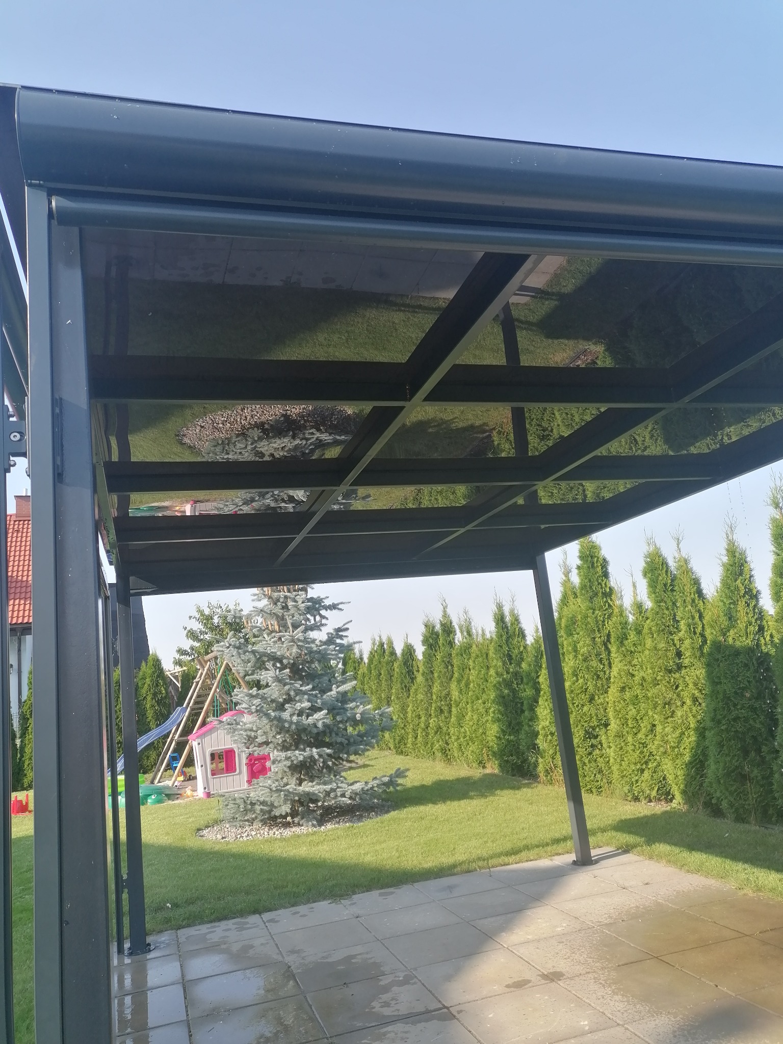 Nowoczesna, grafitowa pergola z metalową konstrukcją i przezroczystym zadaszeniem, w tle zielony ogród z tujami i placem zabaw.