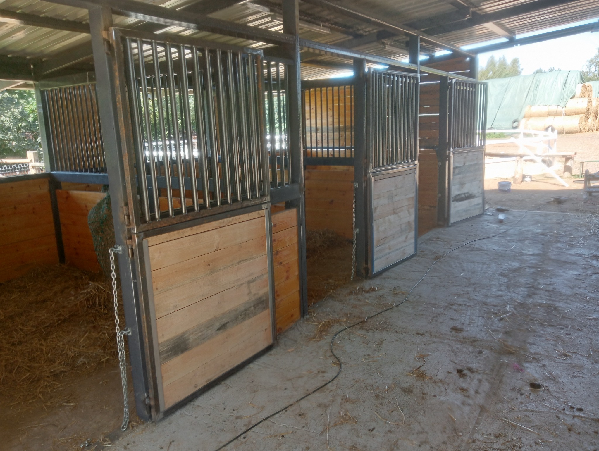 Rząd drewniano-metalowych boksów dla zwierząt gospodarskich, z metalowymi prętami u góry i drewnianymi panelami na dole, wypełnionych słomą, pod zadaszeniem.